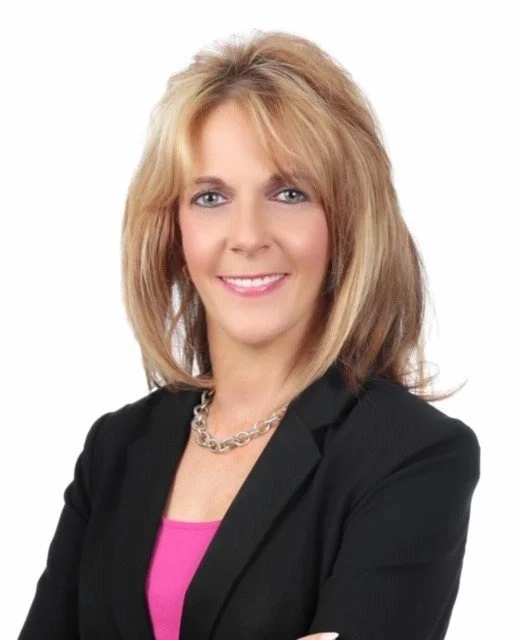 Portrait of a smiling woman with blonde hair, wearing a black blazer and pink top, against a white background.