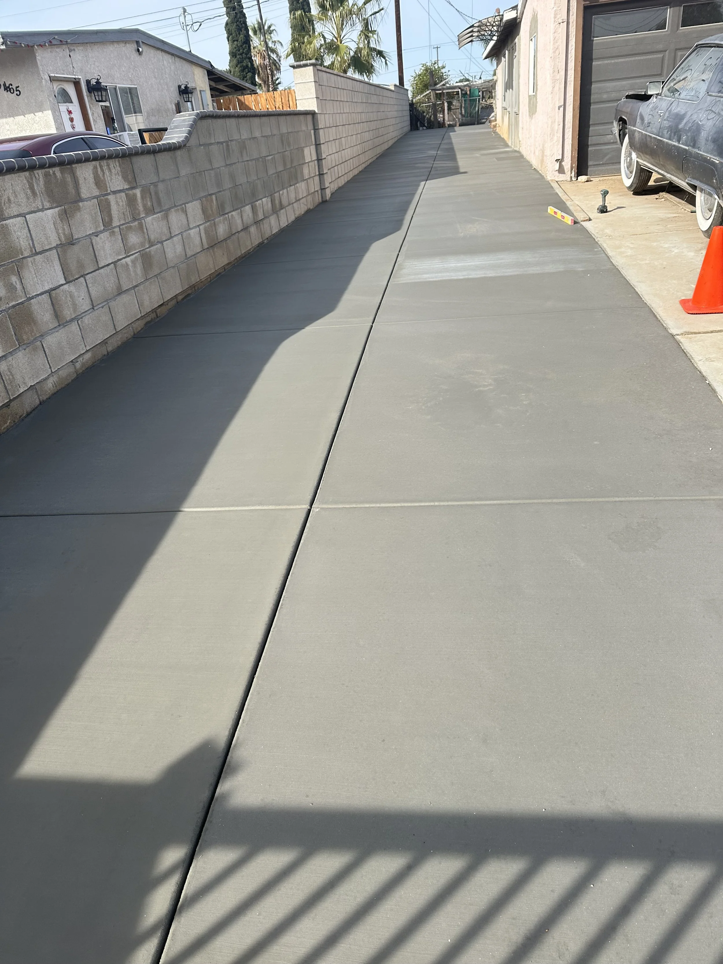 A freshly poured concrete sidewalk next to a brick wall, with construction tools and a parked car nearby in a residential area.