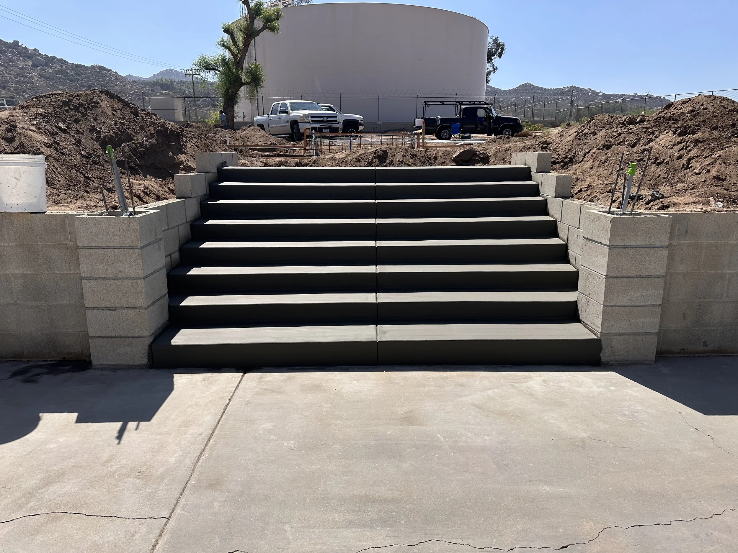 Concrete stairs leading up to a construction site with dirt mounds on both sides, a tree, parked trucks, and mountains in the background.