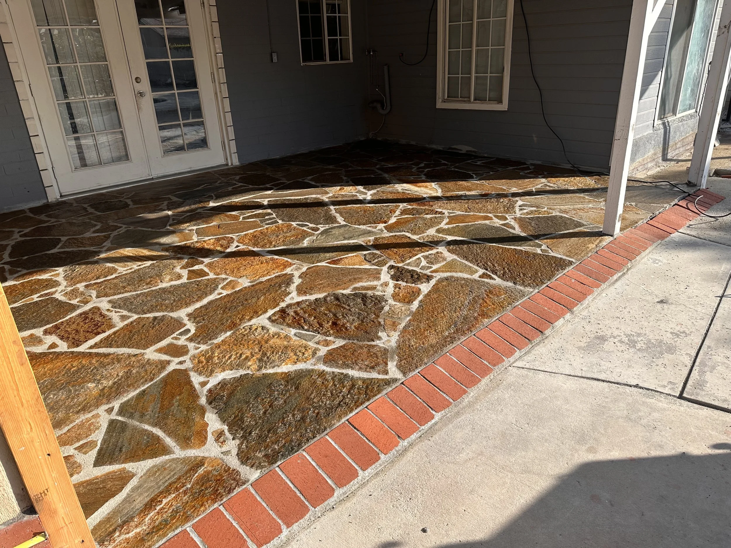 Patio area with a stone floor, a brick border, a gray house wall with a window and a door with glass panes, and some wires hanging on the wall. An area of sunlight and shadow on the floor.