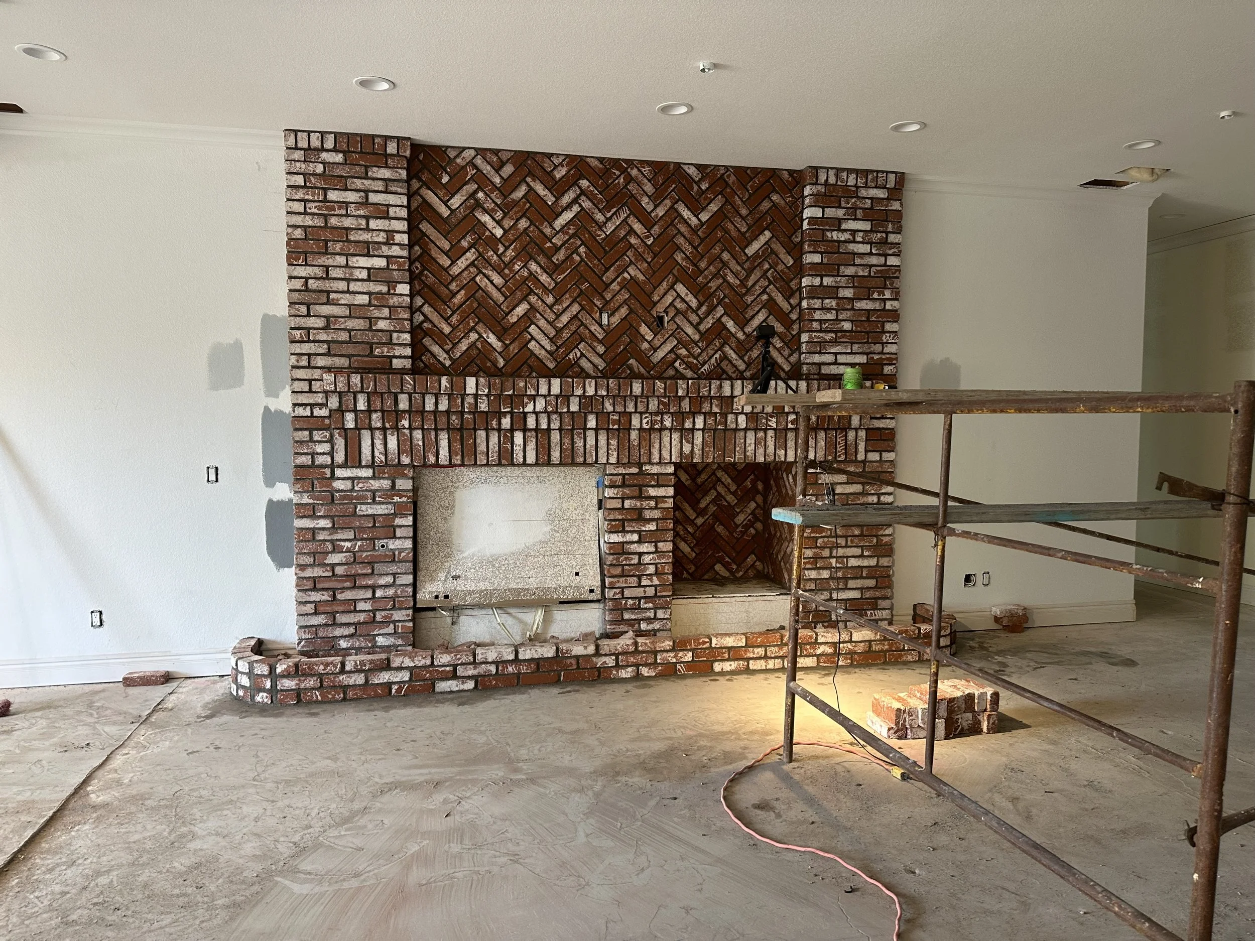 A brick fireplace with a herringbone pattern in a room under construction. The room has unfinished floors, electrical outlets, and a scatter of construction tools and materials.