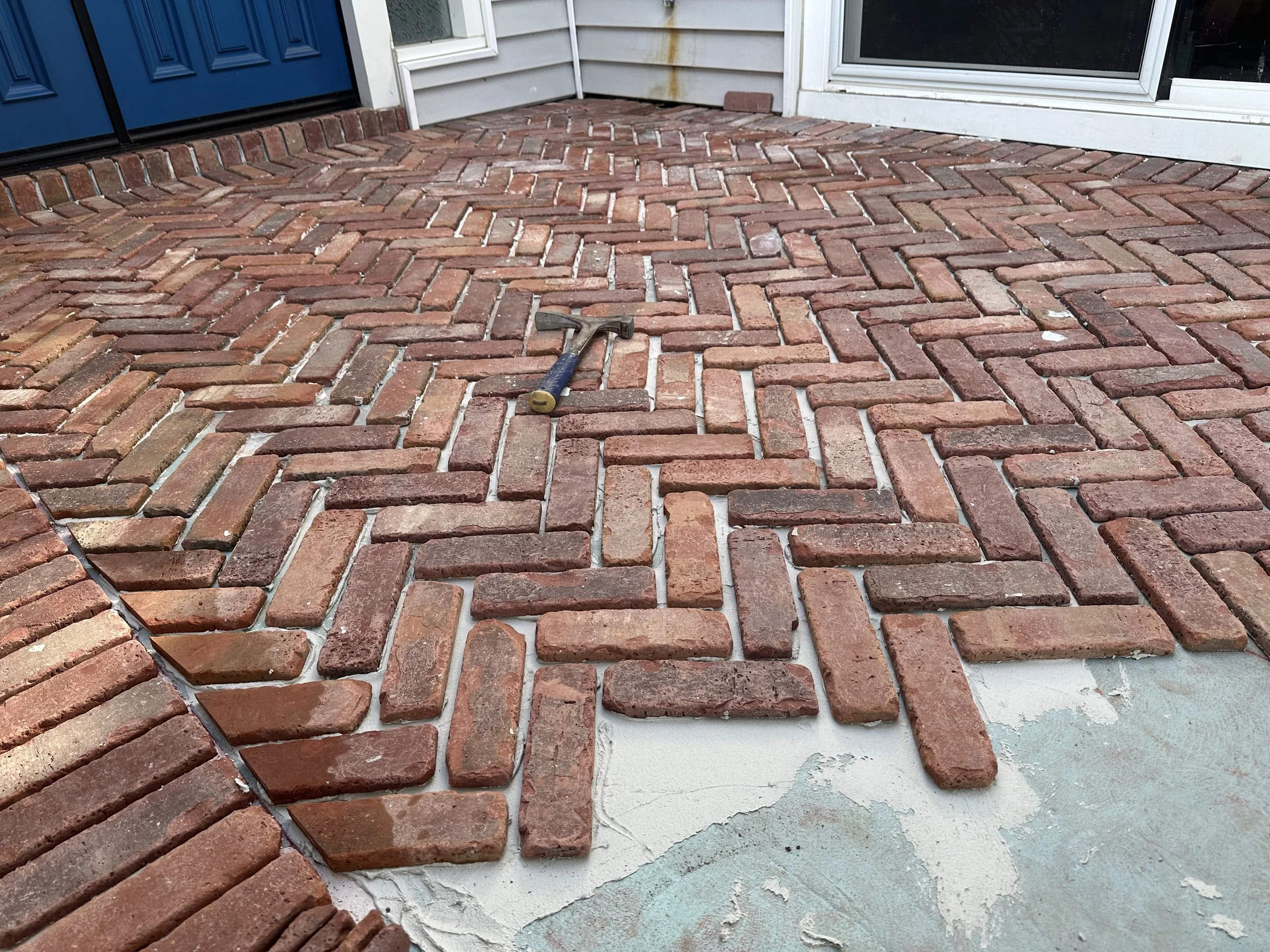 A brick patio with a hammer laying on the bricks near a house entrance, showing a blue door and window.