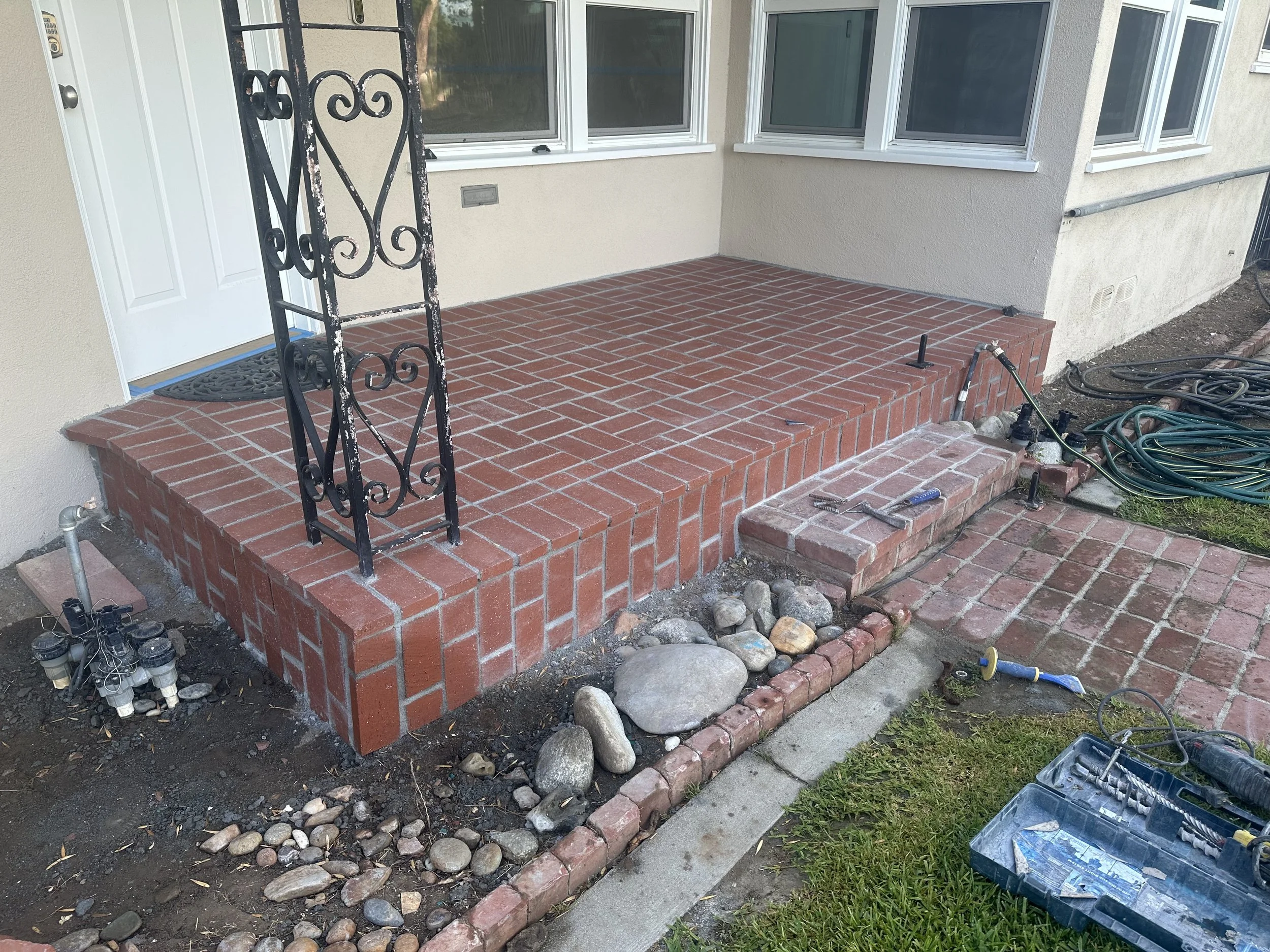 The image shows a brick patio area outside a house under construction or renovation. There is a black decorative iron railing on the left side of the patio, a sliding door with a white frame to the left, and a large window with a white frame on the r