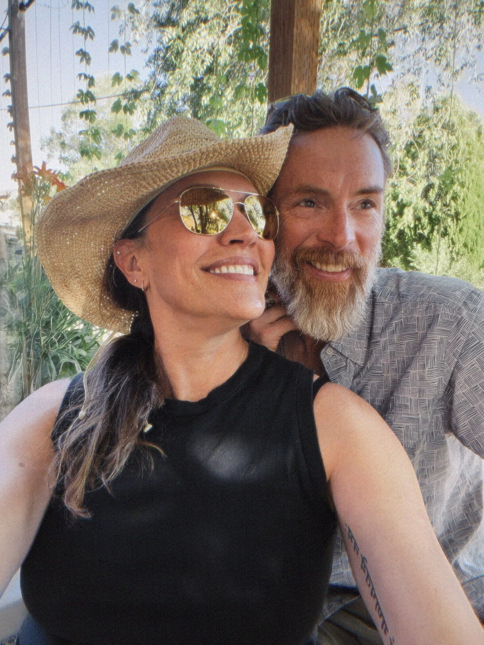 A woman with dark hair, sunglasses, and a straw hat smiling. Next to her is a man with a beard and mustache, also smiling. They are outdoors with trees and greenery in the background.