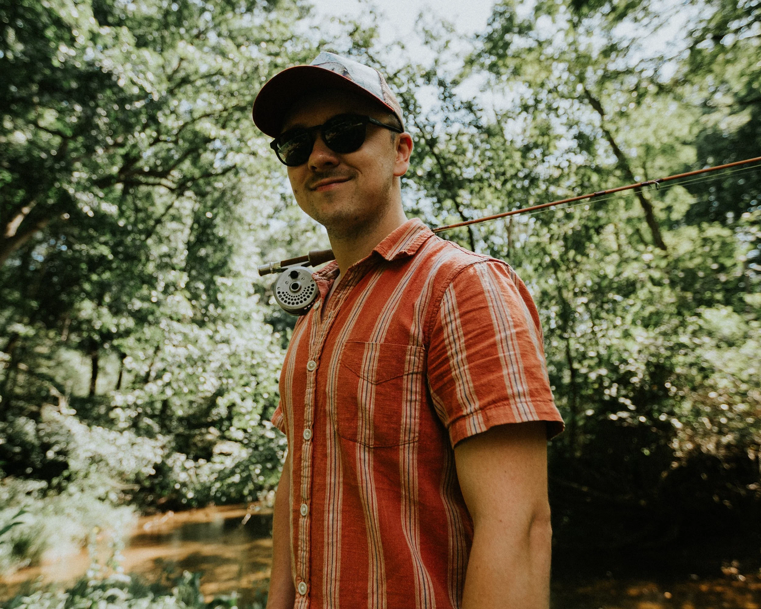 A man wearing sunglasses and a baseball cap with a plaid pattern, smiling, in an outdoor setting with trees and sunlight in the background. He is dressed in a short-sleeved, striped, orange and white button-up shirt.