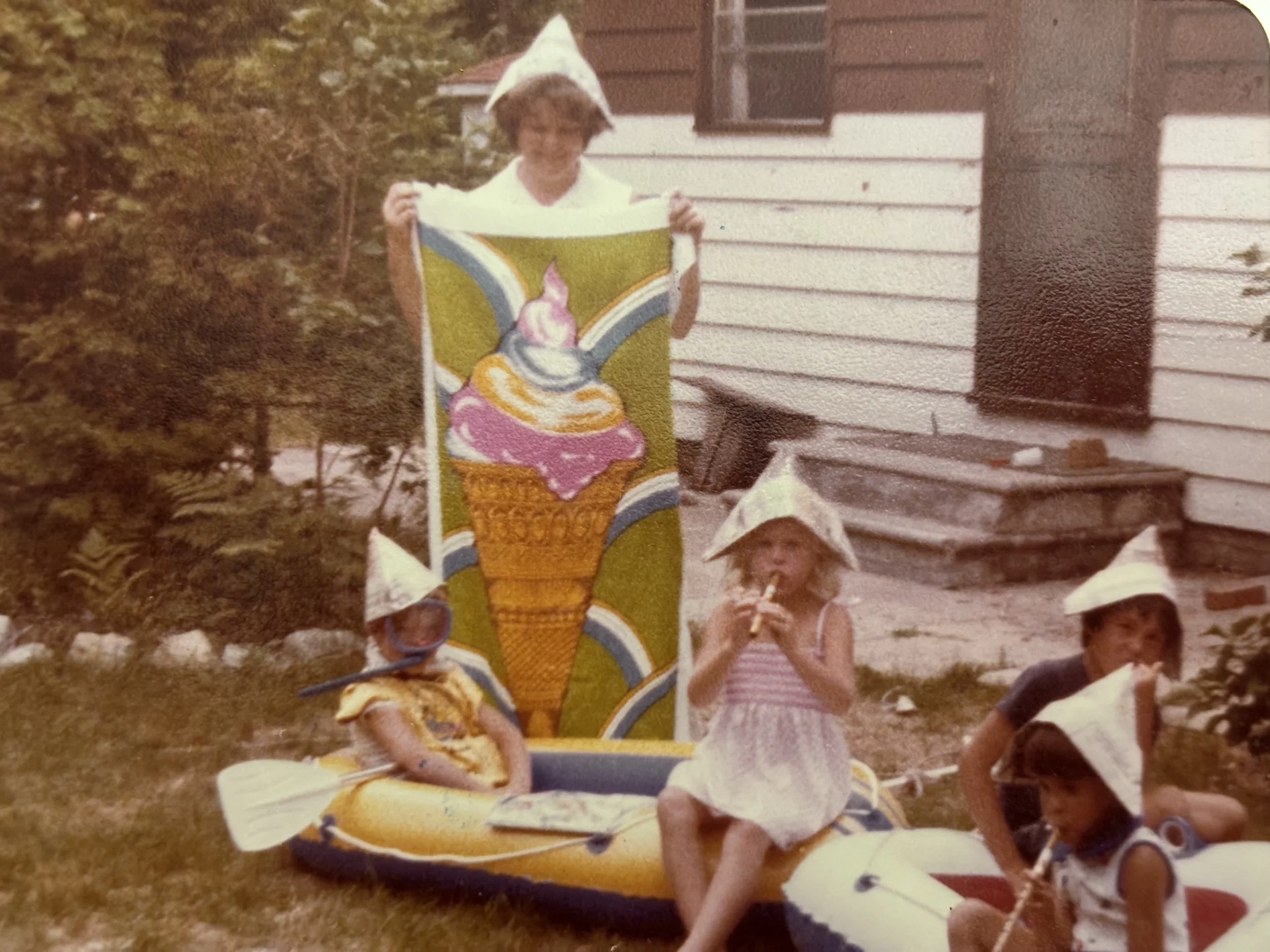 A young boy dressed as an elf standing outdoors during daytime, holding a colorful sign with a bunny and cupcake design, with other children nearby and a house with horizontal siding in the background.