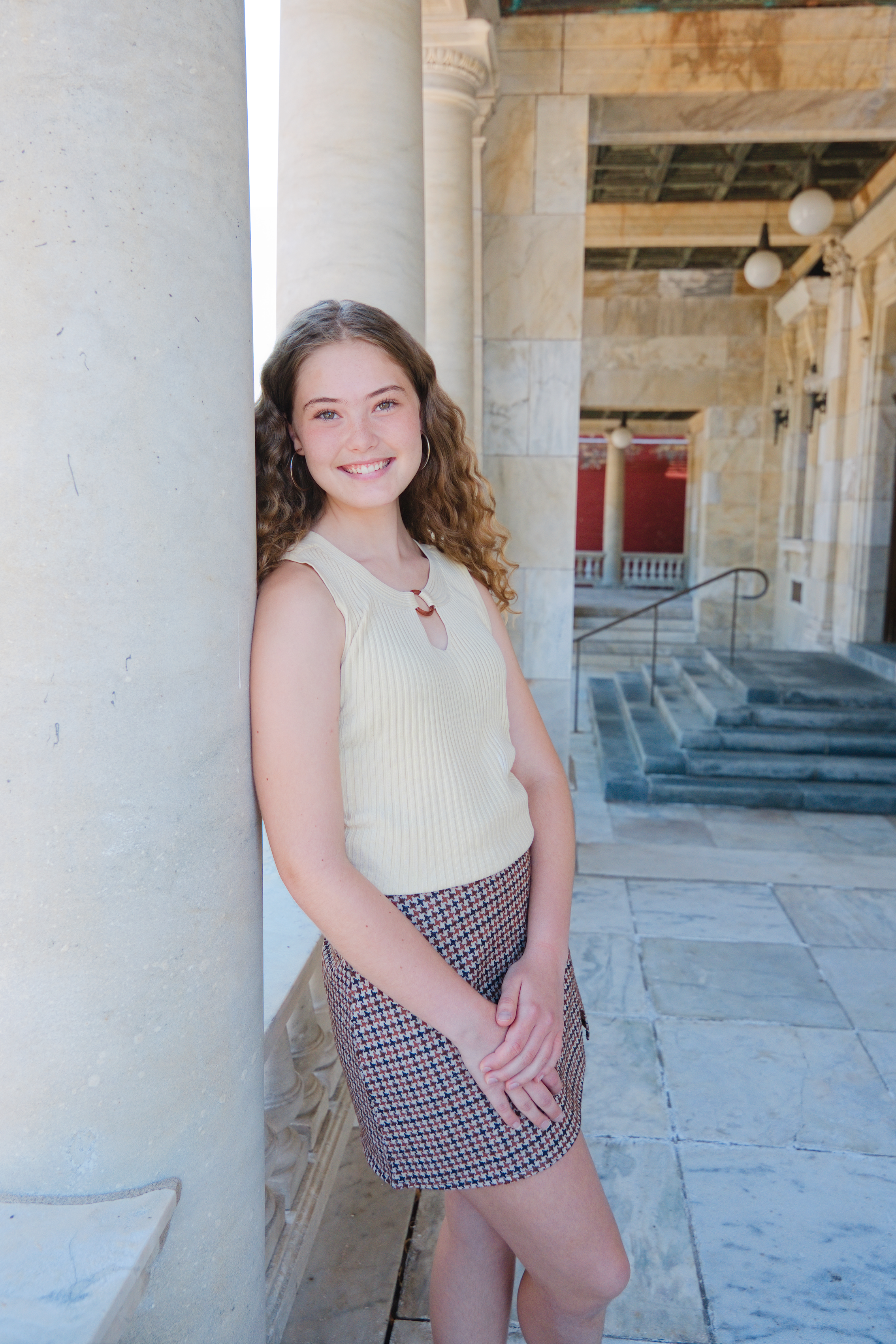 Young woman with curly hair smiling, leaning against a column in front of a classical-style building with marble stairs and columns.