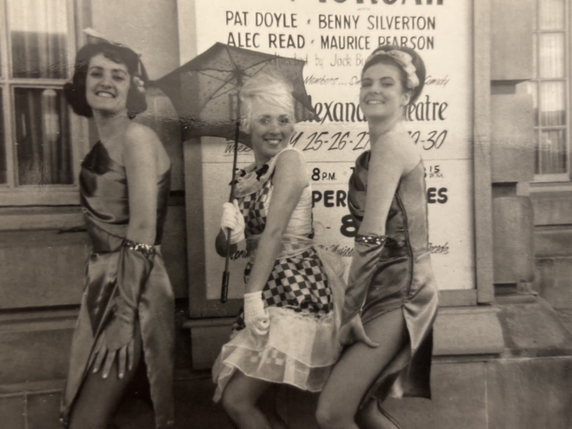 Three women in vintage attire standing in front of a poster, one holding an umbrella, smiling for the photo.