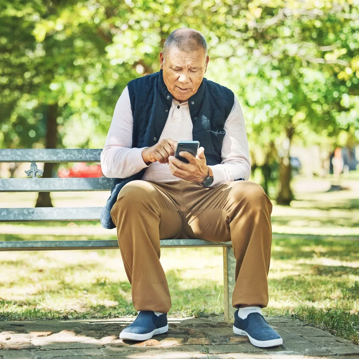 An elderly man sitting on a park bench, looking at his smartphone, surrounded by trees and greenery.