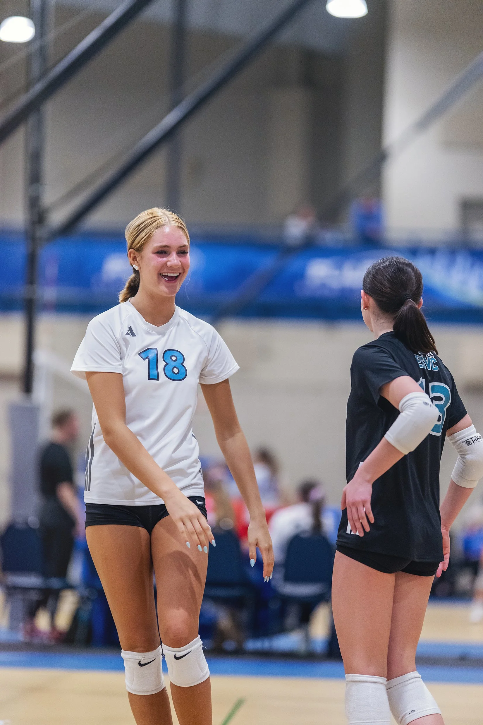 Two female indoor volleyball players standing and talking on the court, one wearing a white jersey with the number 18 and the other wearing a black jersey with the number 12. The player in white is smiling.