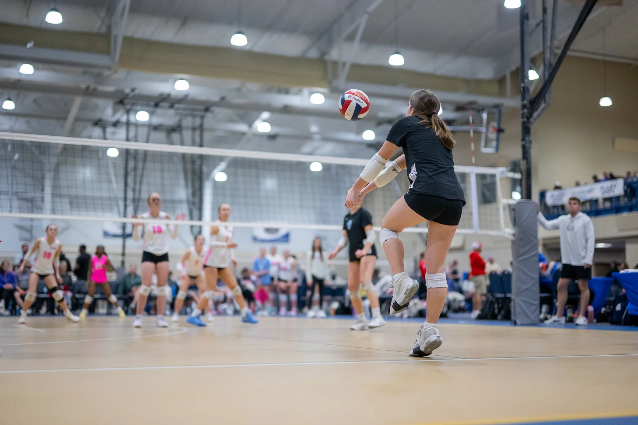 A female volleyball player in black sportswear hitting the ball during a match inside a gymnasium, with other players and spectators watching in the background.