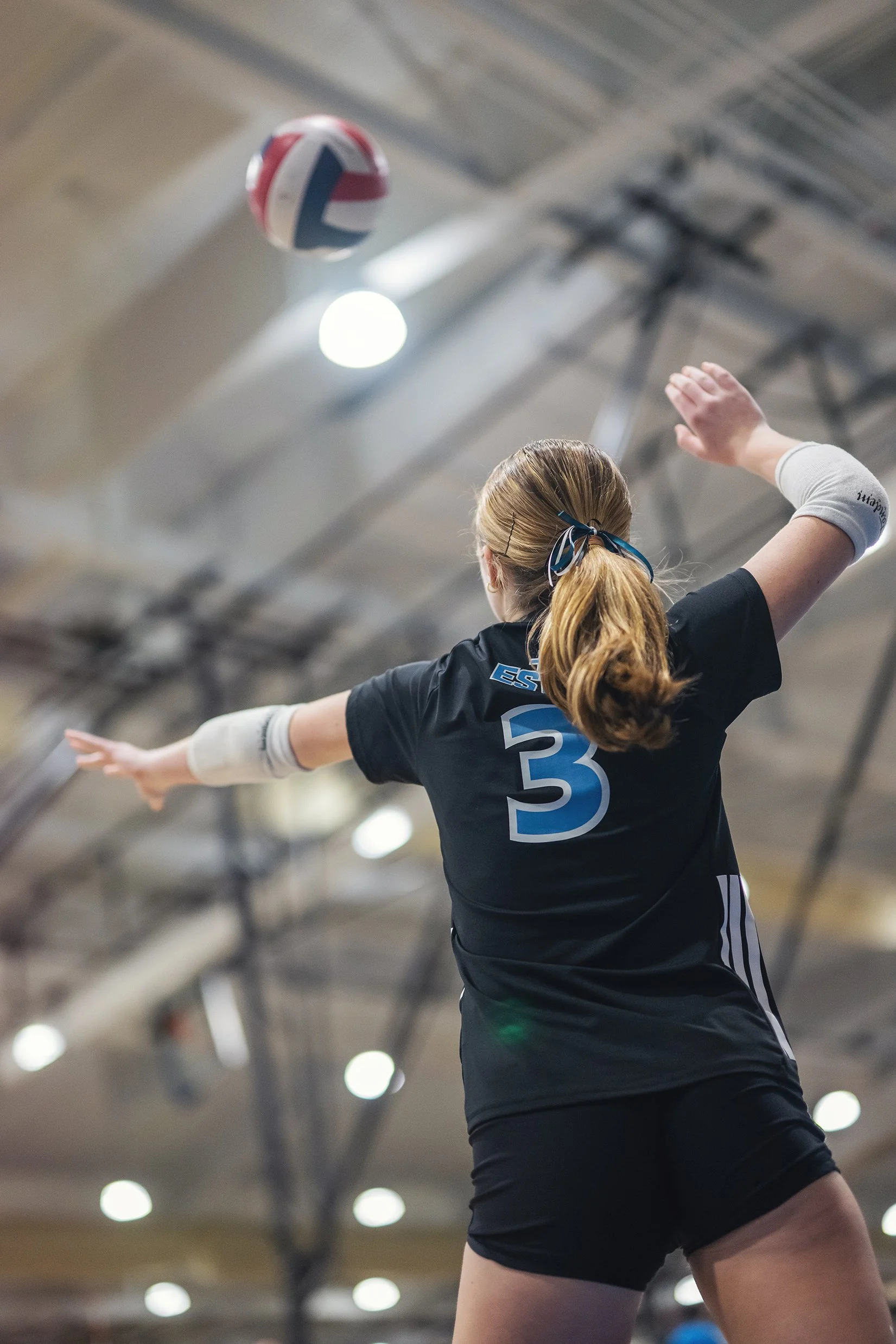 A female volleyball player with blonde hair tied in a ponytail, wearing a dark sports jersey with the number 3, is reaching out to hit a volleyball in an indoor gymnasium.