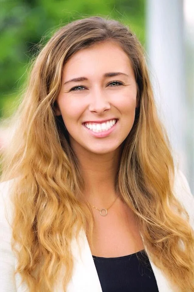 Smiling young woman with long wavy blonde hair, wearing a white blazer and black top, outdoors with greenery in the background.