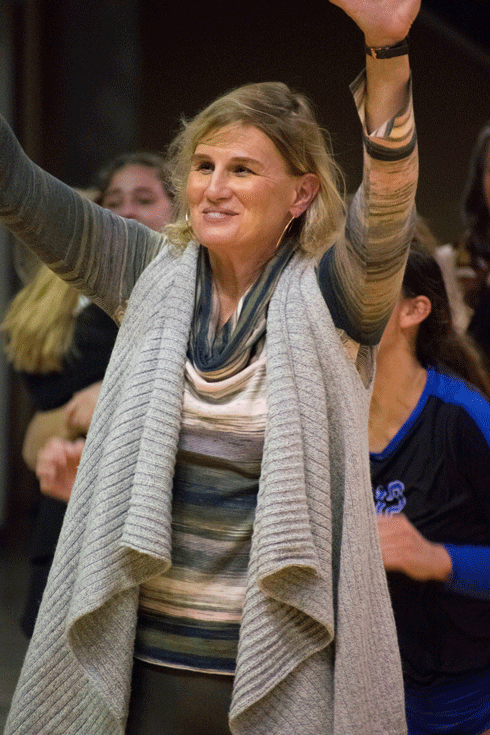 A woman with blonde hair smiling and raising her hand in a crowd. She is wearing a striped shirt and a gray scarf.