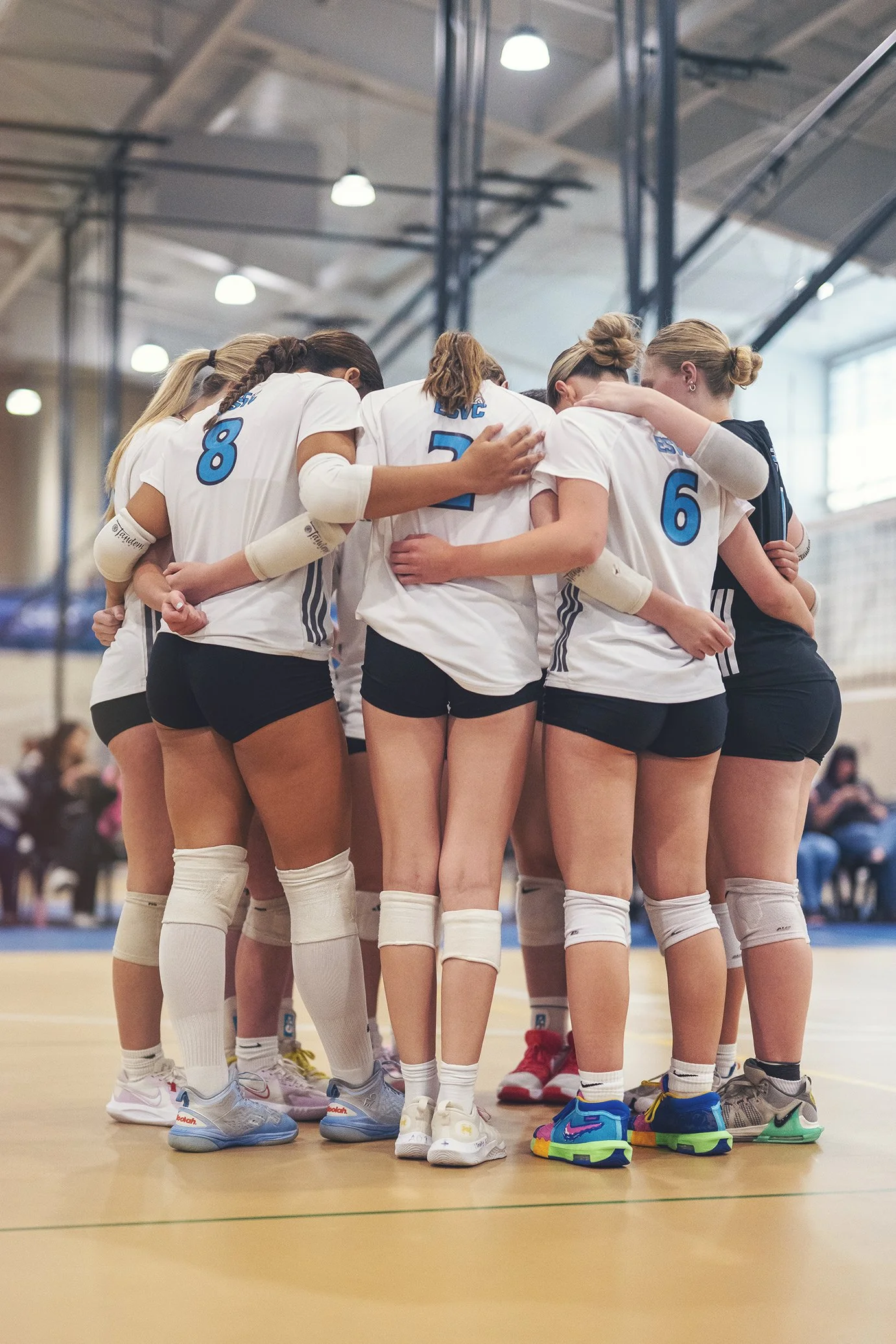 A group of young female volleyball players standing in a circle with their arms around each other, wearing white uniforms with blue numbers, inside a gymnasium.