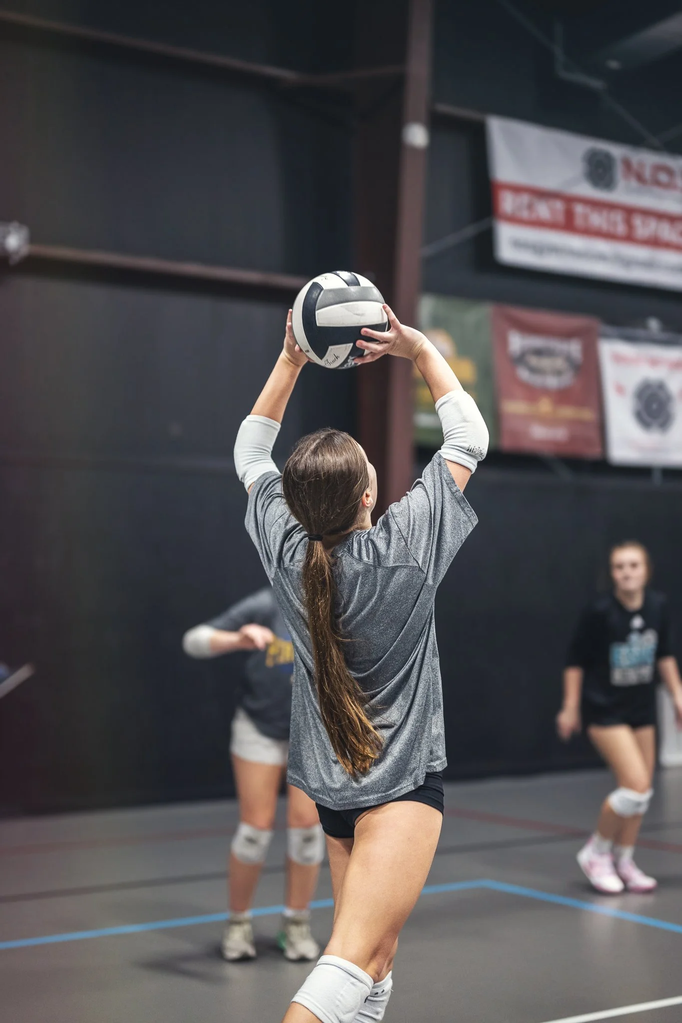 A young girl in a gray sports shirt and black shorts is hitting a volleyball in an indoor gym. Other children are visible in the background, some standing and watching.