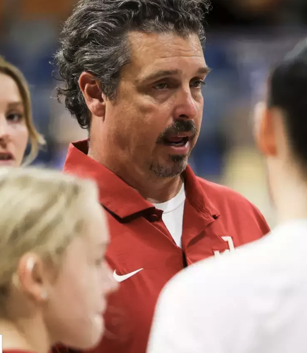 A man with dark, curly hair, a beard, and a mustache, speaking passionately to a group of people. He is wearing a red sports jacket with a white Nike logo and appears to be in a sports setting.