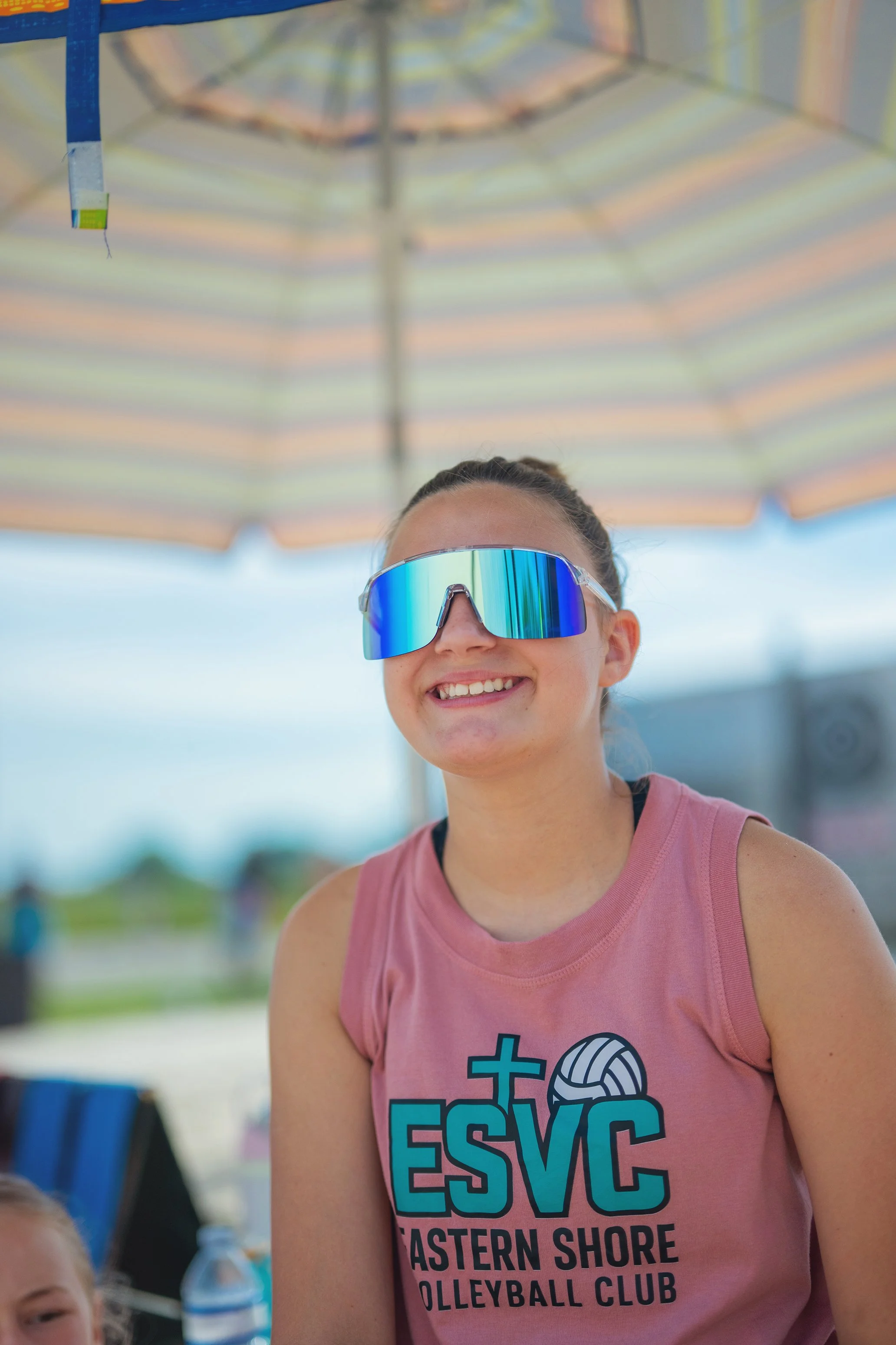 A young woman wearing reflective sports sunglasses and a pink sleeveless volleyball shirt with text, smiling under a large striped beach umbrella.