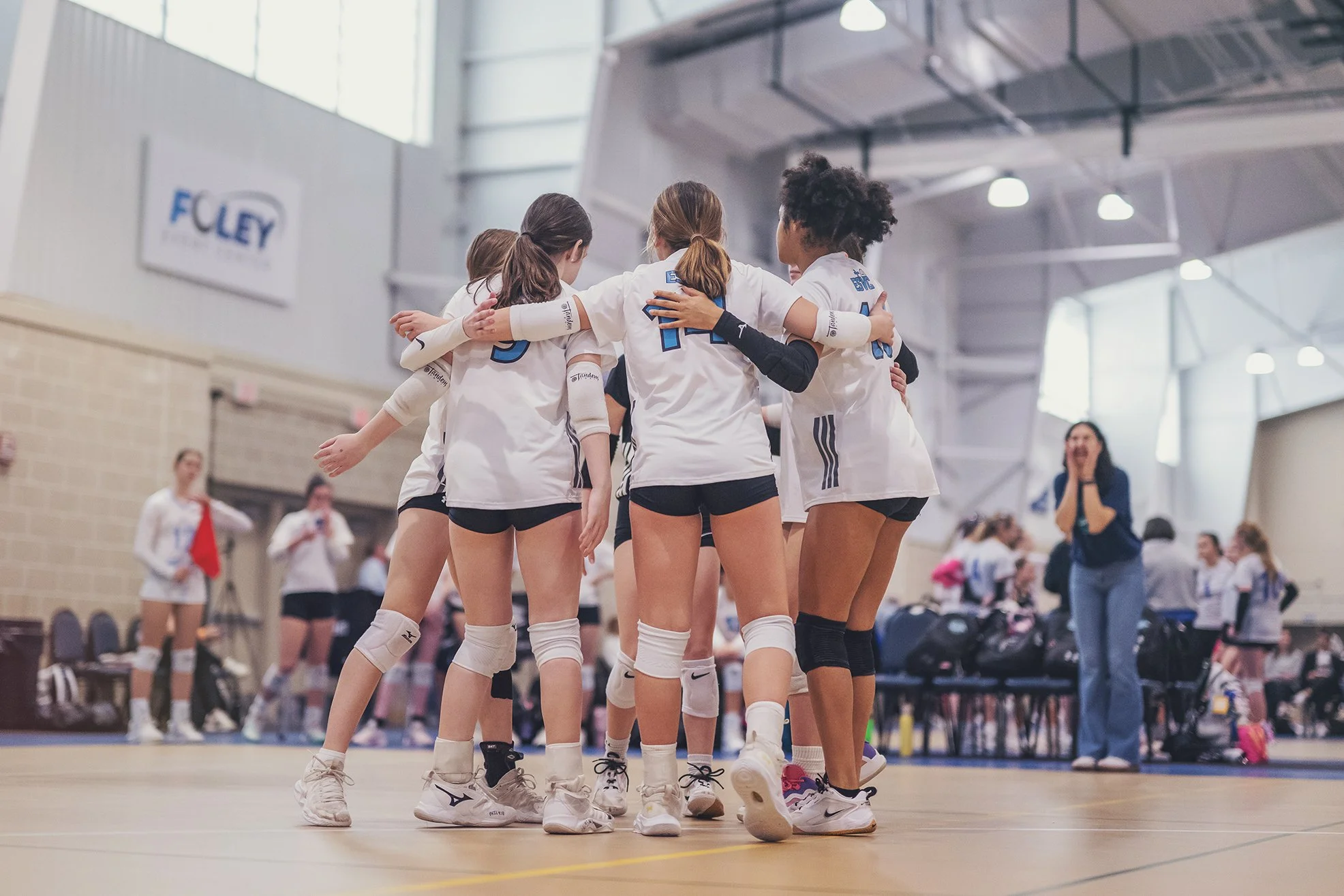 A group of female volleyball players in white uniforms huddle together on an indoor court during a game or practice. Spectators and a coach or referee can be seen in the background.