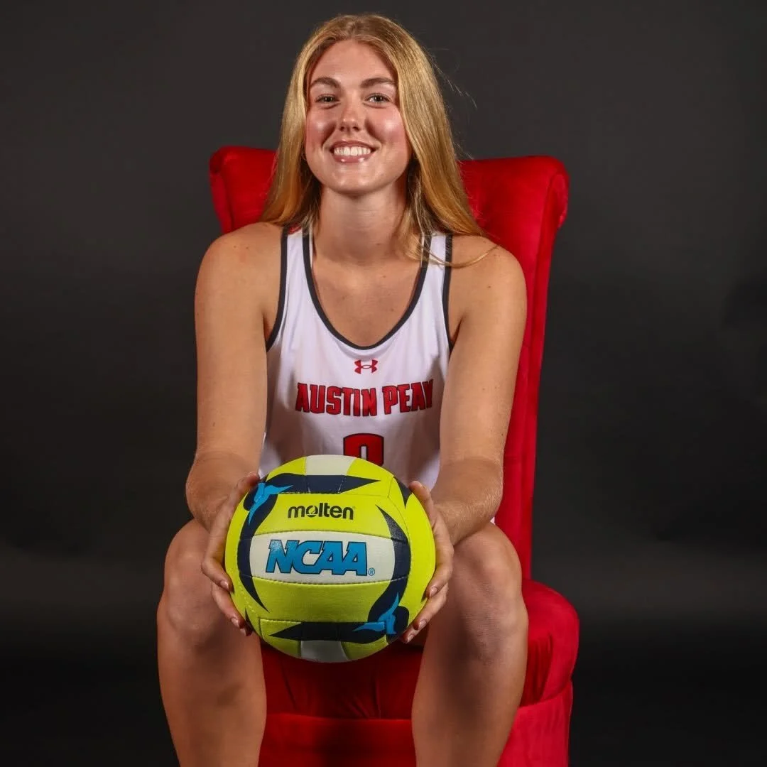A young woman with long blonde hair, wearing a white sports jersey with red accents and 'AUSTIN PEAY' written on it, sitting on a red chair against a black background, holding a yellow NCAA volleyball, smiling at the camera.