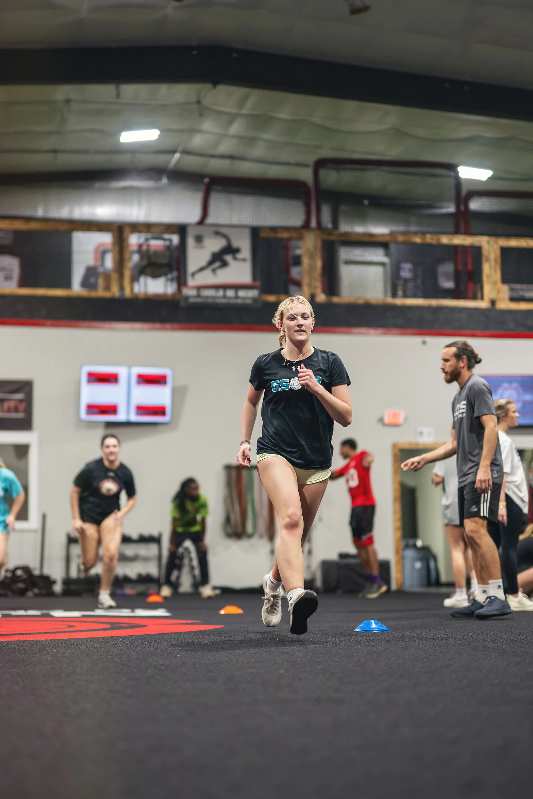 A young woman running on a gym floor during a workout or training session, with several other people and gym equipment in the background.