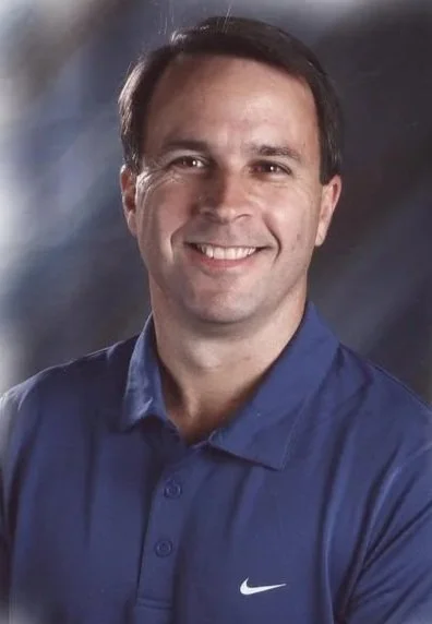 Portrait of a man with short dark hair, smiling, wearing a blue Nike polo shirt, against a dark, blurred background.