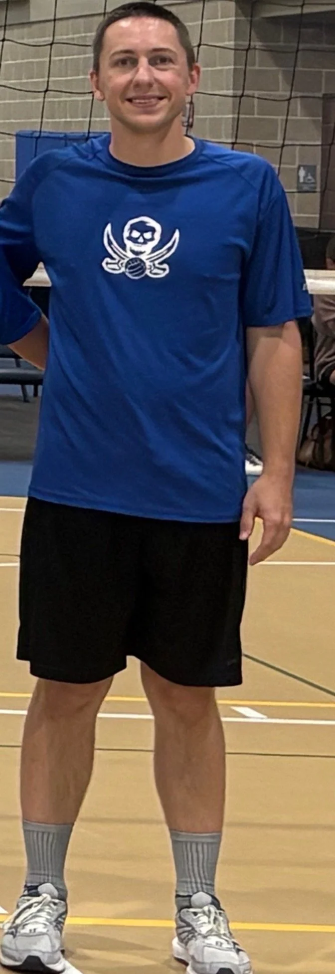 A man standing in a gymnasium wearing a blue sports shirt with a skull and wings logo, black shorts, gray socks, and running shoes.