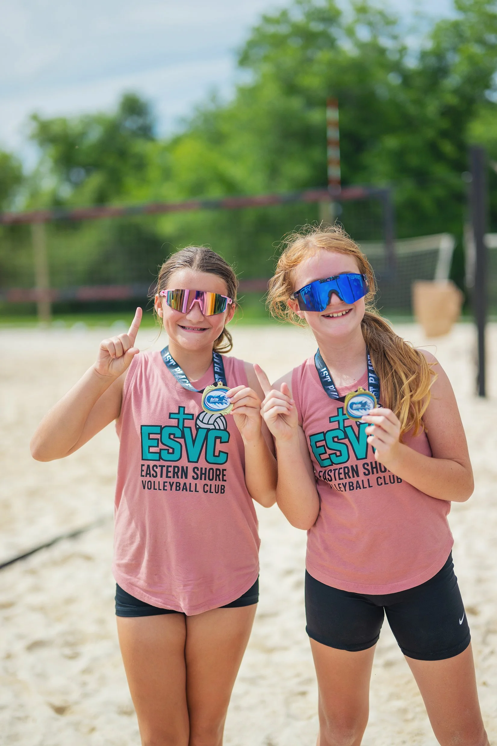 Two young girls standing side by side on a beach volleyball court, wearing pink shirts with the text 'ESVC Eastern Shore Volleyball Club', sunglasses, and medals around their necks, smiling and holding up one finger each.