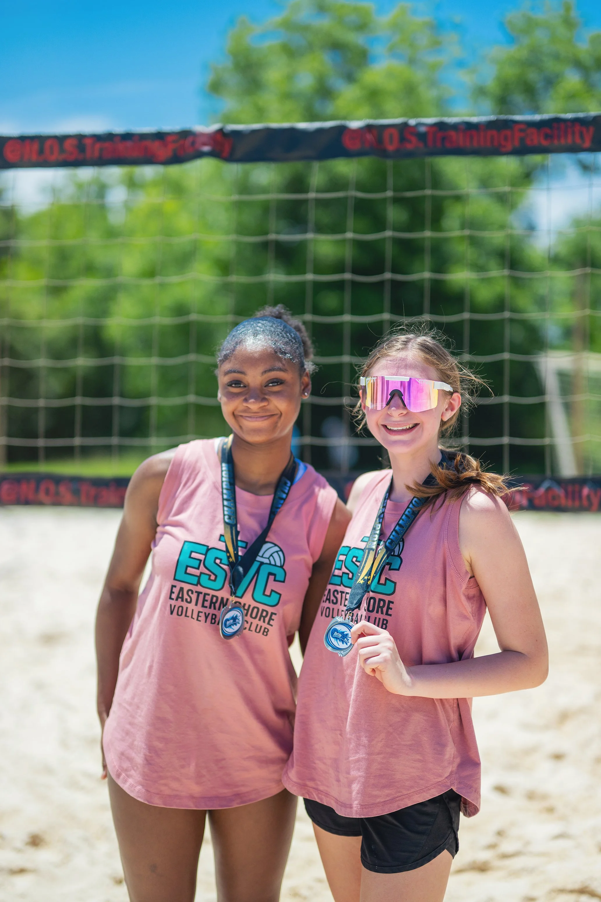 Two young girls standing on a beach volleyball court holding medals, wearing pink sleeveless sport jerseys with "Eastern Shore Volleyball Club" printed on them, smiling at the camera, with a volleyball net behind them and a blue sky with green trees in the background.