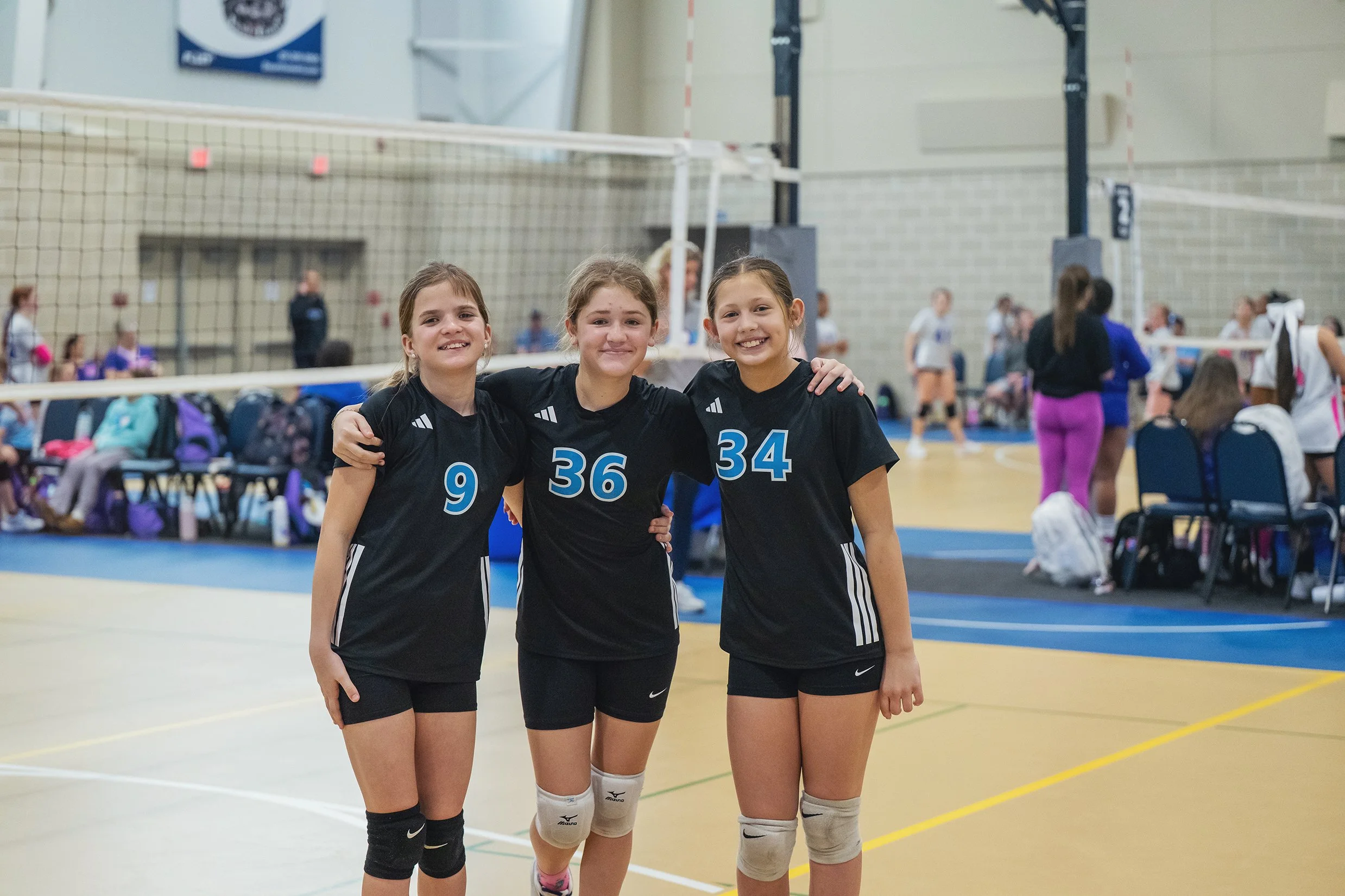 Three young female volleyball players in black uniforms with blue numbers stand on an indoor volleyball court, smiling and posing for a photo with their arms around each other.