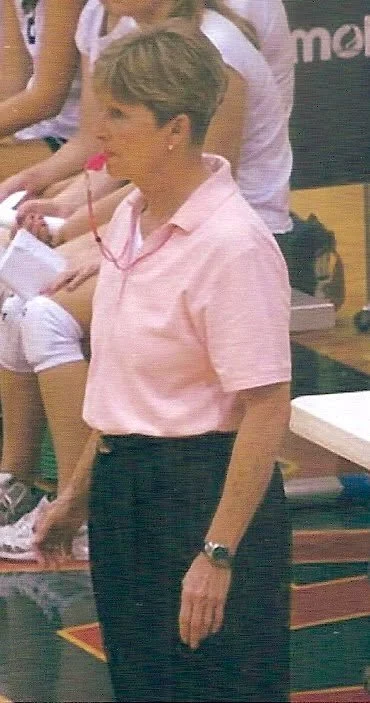 A woman with short blonde hair wearing a light pink polo shirt and black pants stands indoors. She has a watch on her left wrist and appears to be looking to her left.