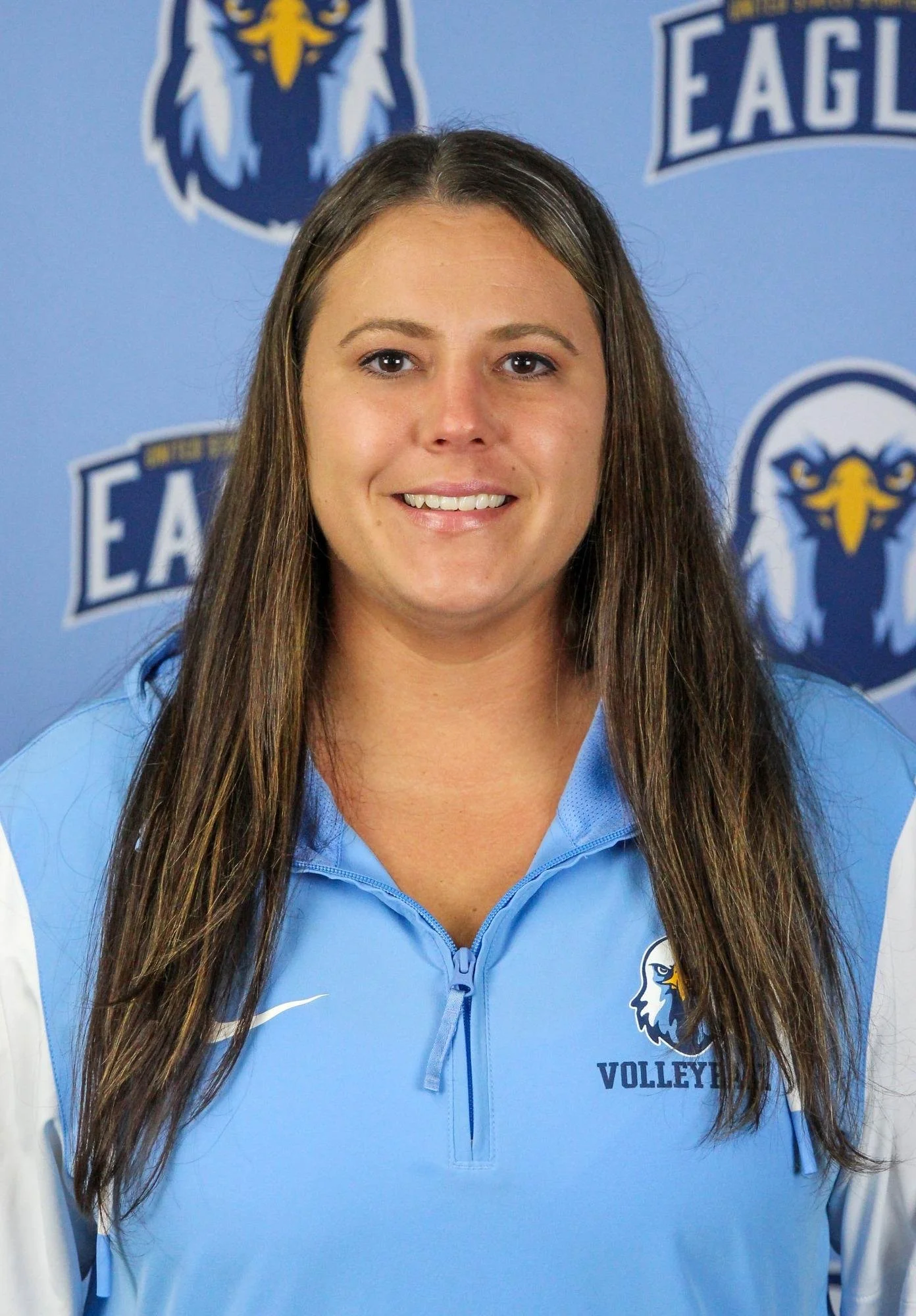 A woman with long brown hair wearing a light blue volleyball jacket standing in front of a blue background with logos and text related to volleyball.