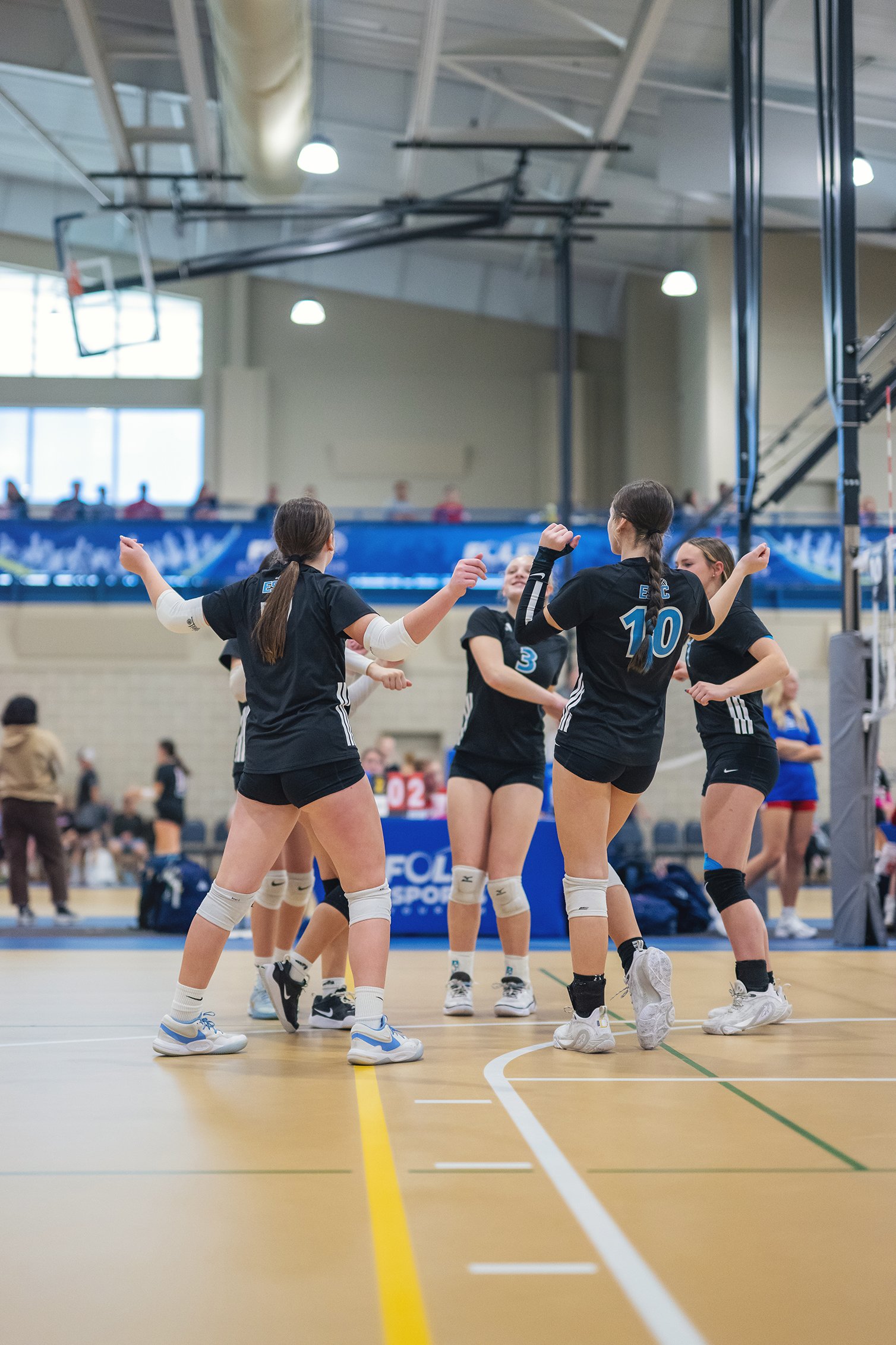 A women's volleyball team huddles together on an indoor court during a game or practice, with some players smiling and raising their arms. The gym has bright lights, a basketball hoop, and fans or spectators in the background.