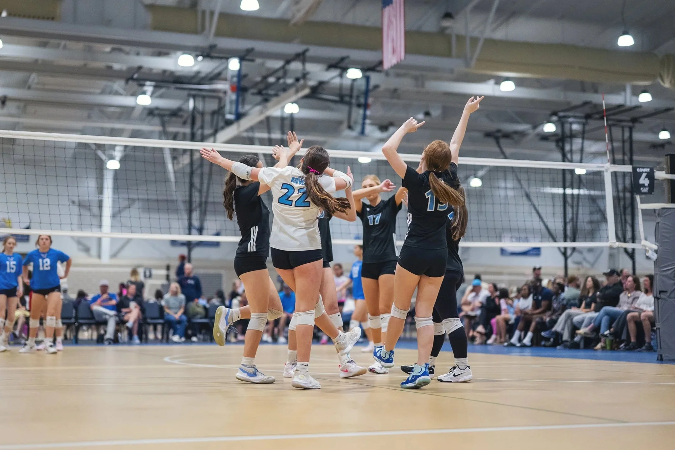 Girls volleyball team celebrating on the court during a match in an indoor gymnasium with spectators watching.