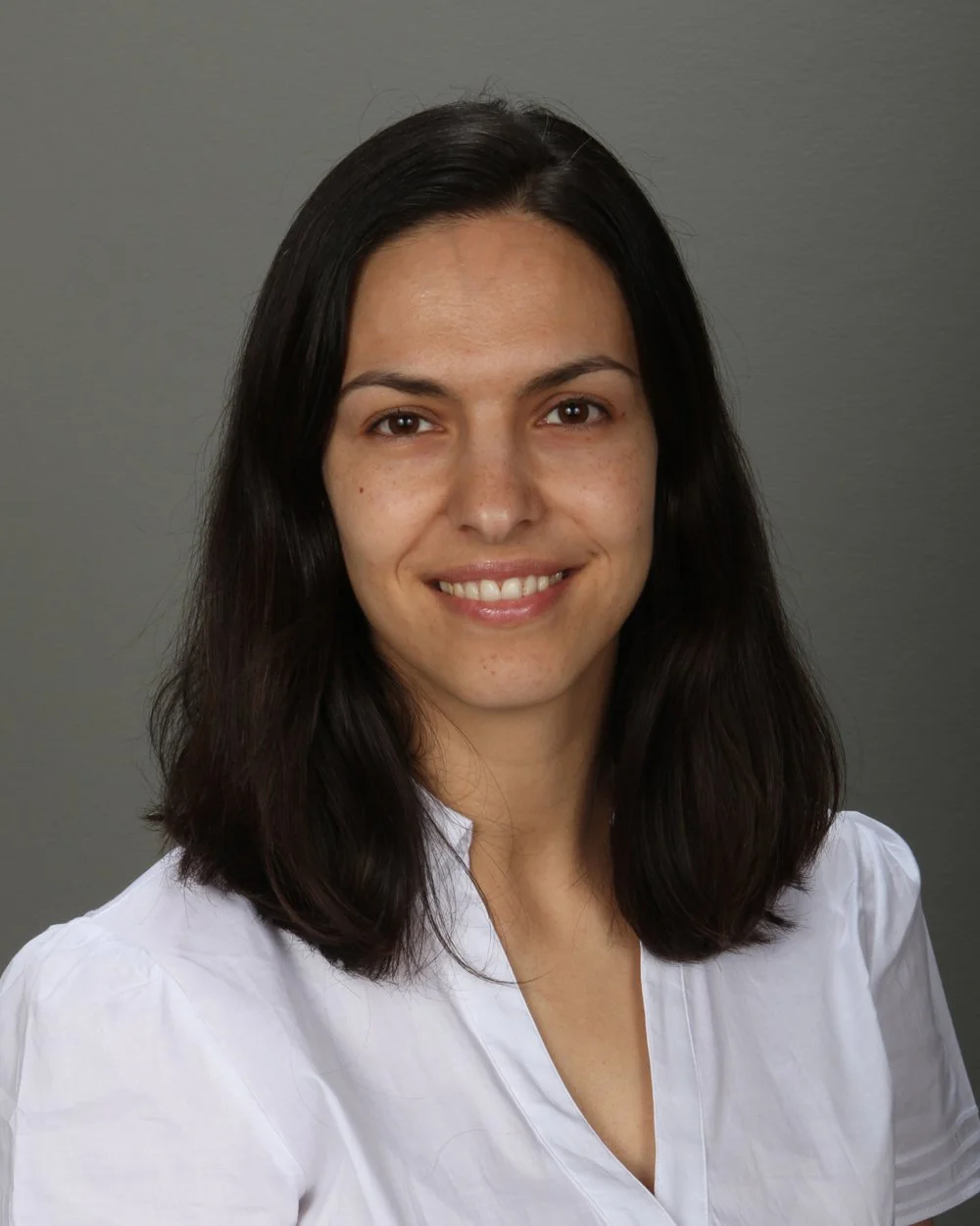 Portrait of a woman with shoulder-length dark hair, smiling, wearing a white blouse against a gray background.