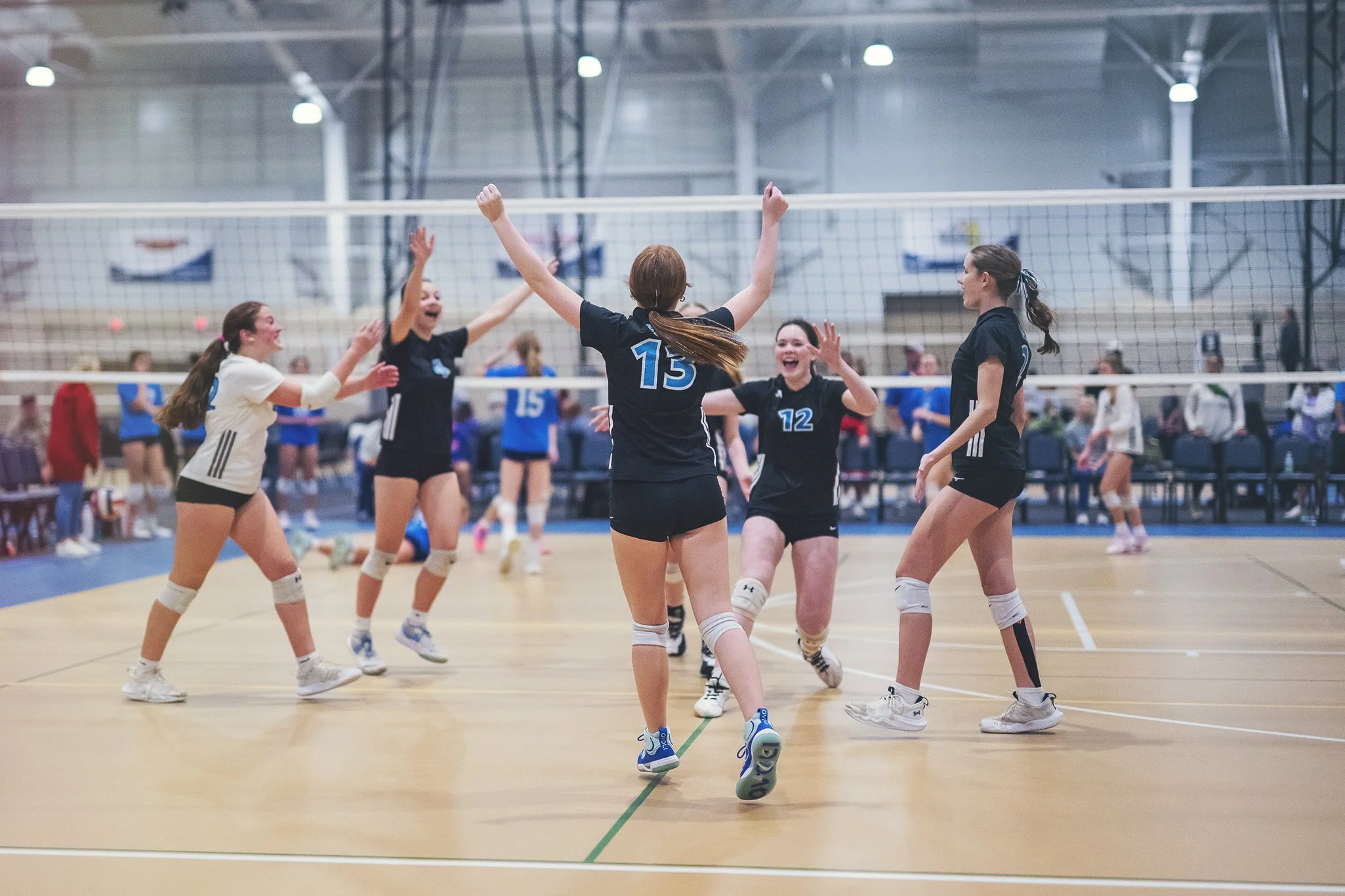 Young female volleyball players celebrate on an indoor court, with some jumping and others smiling, wearing black and white jerseys, under bright gym lights.