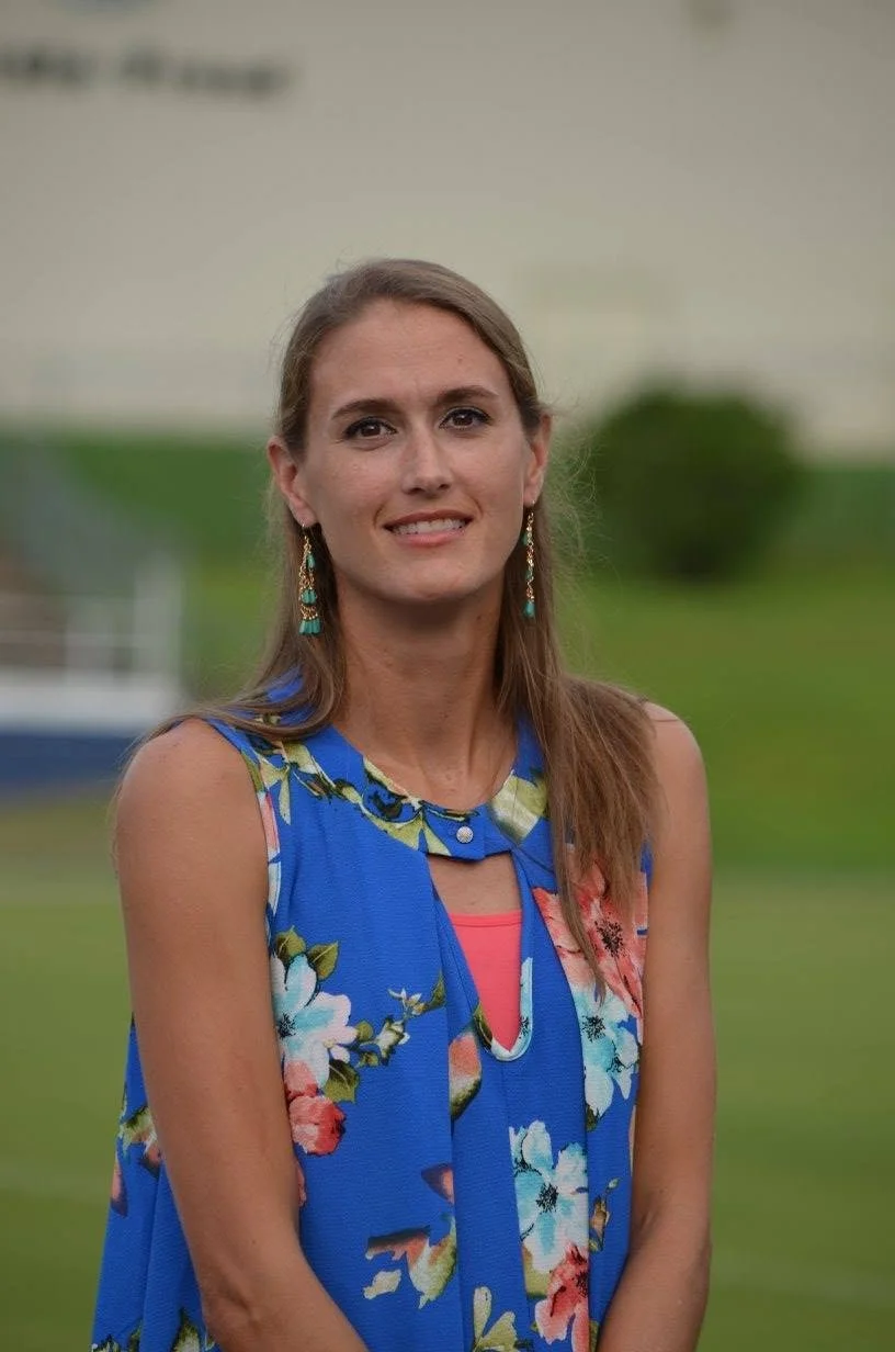 A woman with long brown hair wearing a blue floral sleeveless top, gold earrings, standing outdoors with blurred green background.