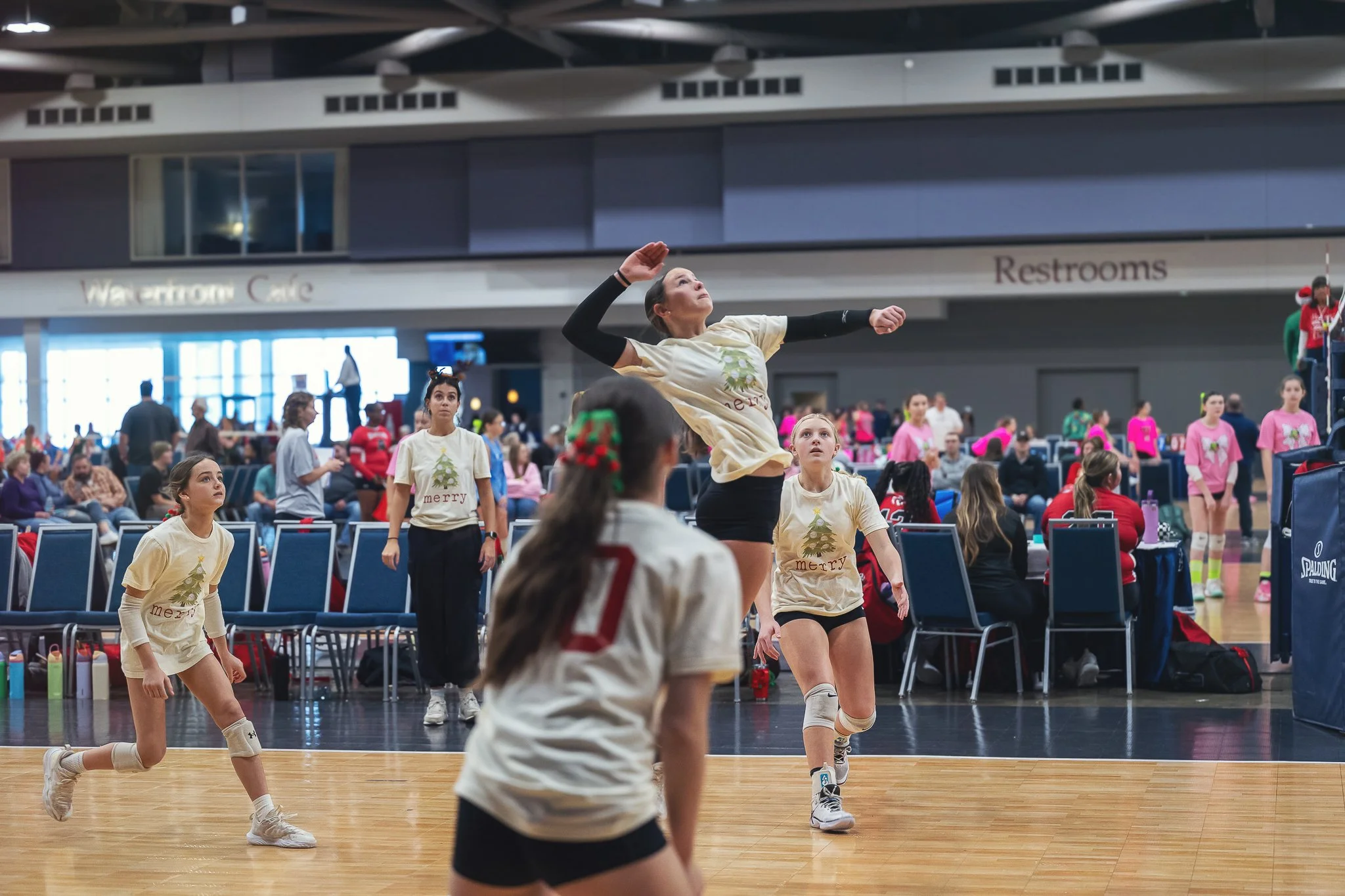 A young woman playing volleyball indoors at a sports event, surrounded by other players and spectators.