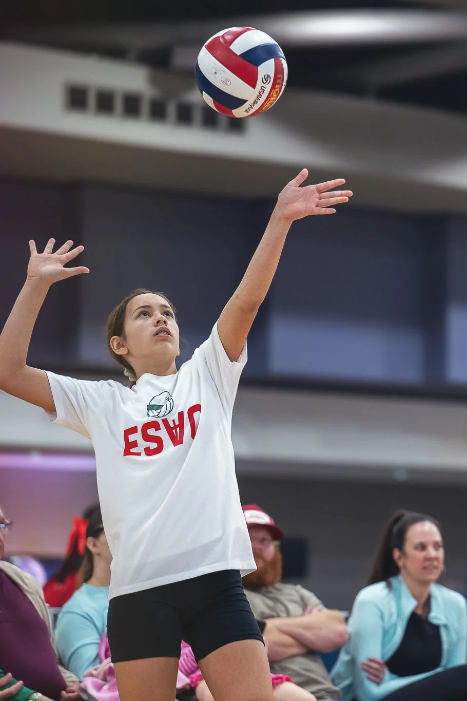 A young girl playing volleyball indoors, preparing to serve, wearing a white t-shirt with red lettering and black shorts, with spectators watching in the background.