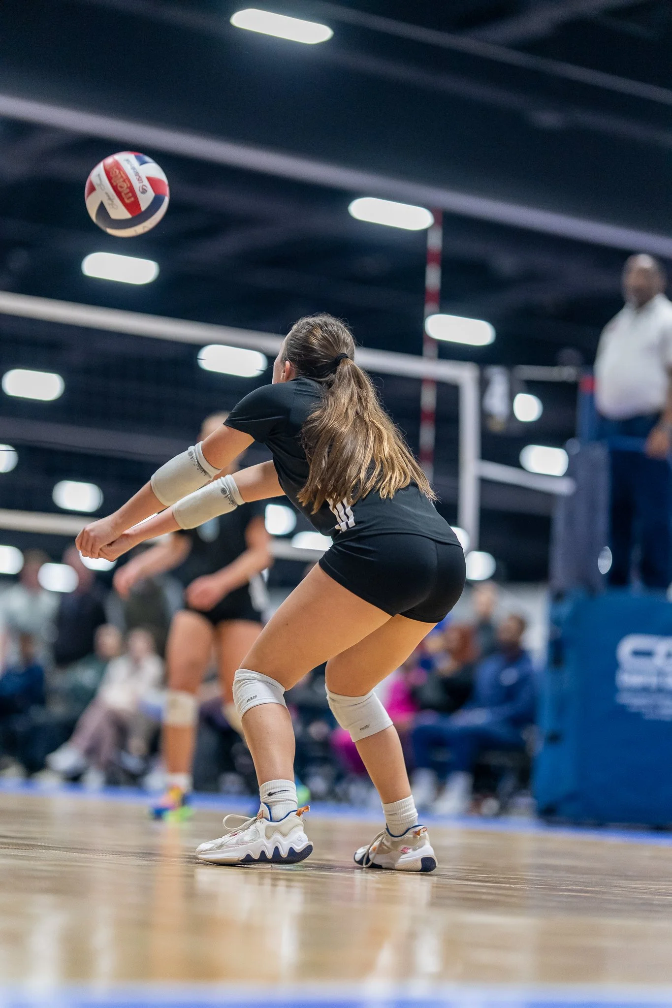 A female volleyball player in black athletic gear and white knee pads is positioned to receive a volleyball during a game in an indoor gymnasium with spectators and officials in the background.