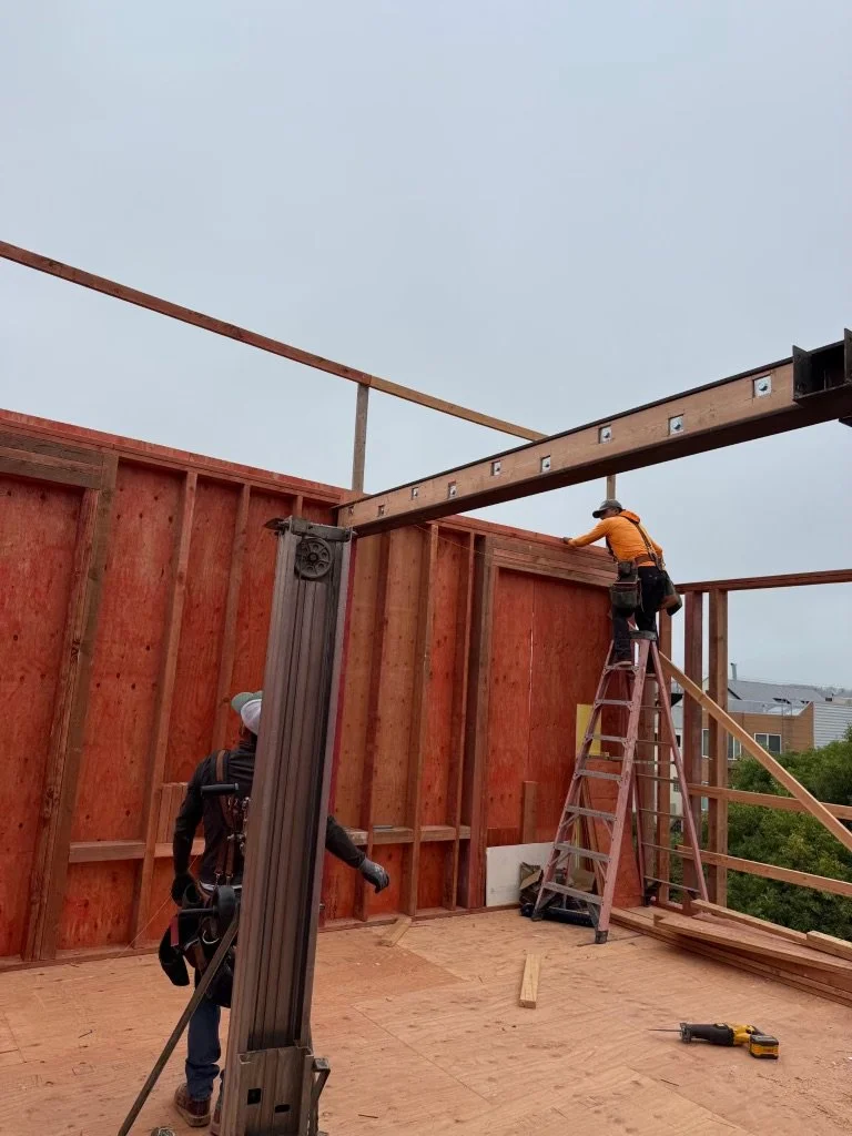 Construction workers are building a wooden structure, with one worker standing on a ladder securing a beam, and another worker standing on the floor adjusting materials; tools and supplies are visible on the floor.