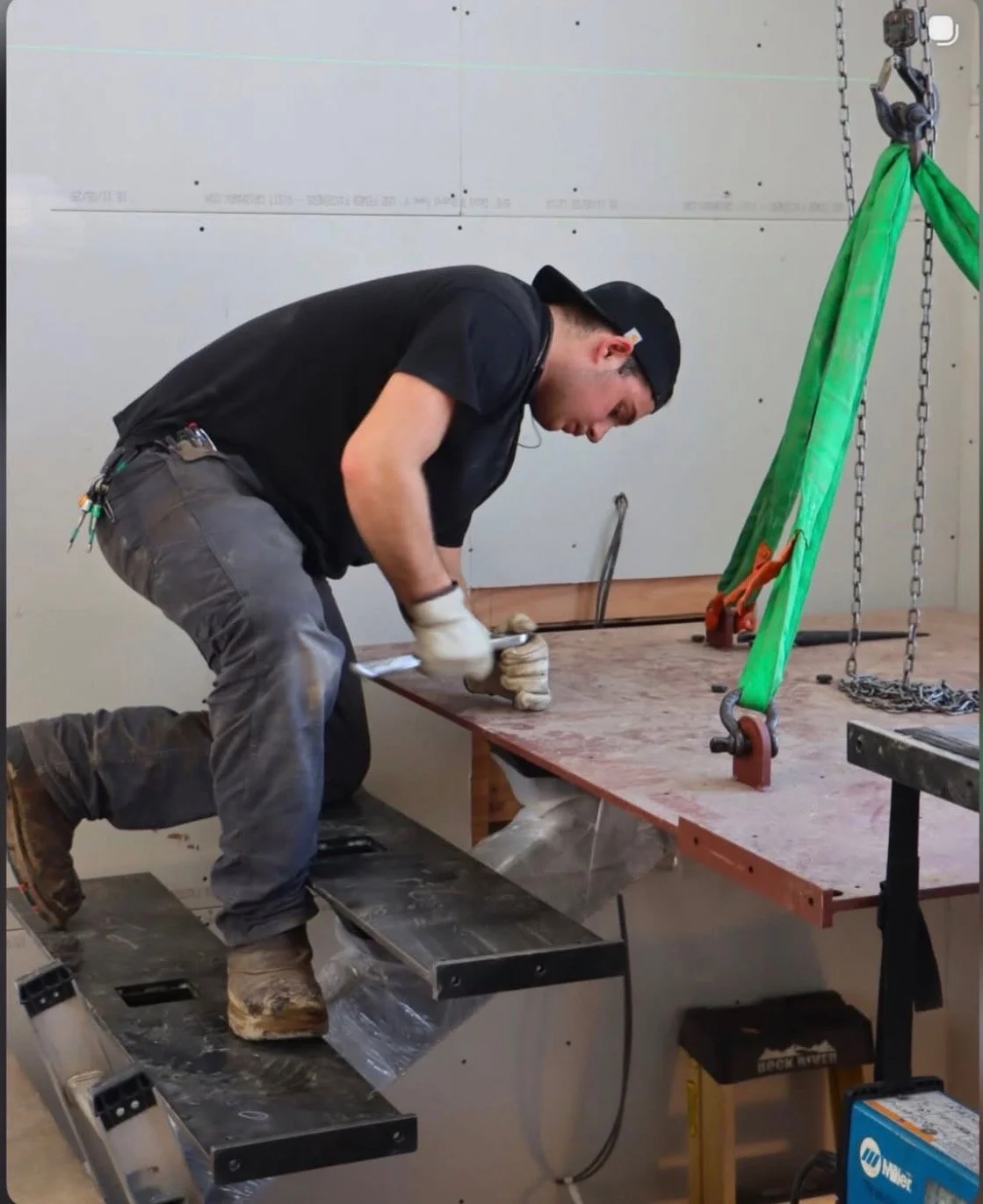 A man kneeling on a black platform working on a wooden workbench with a green pulley system attached to chains.