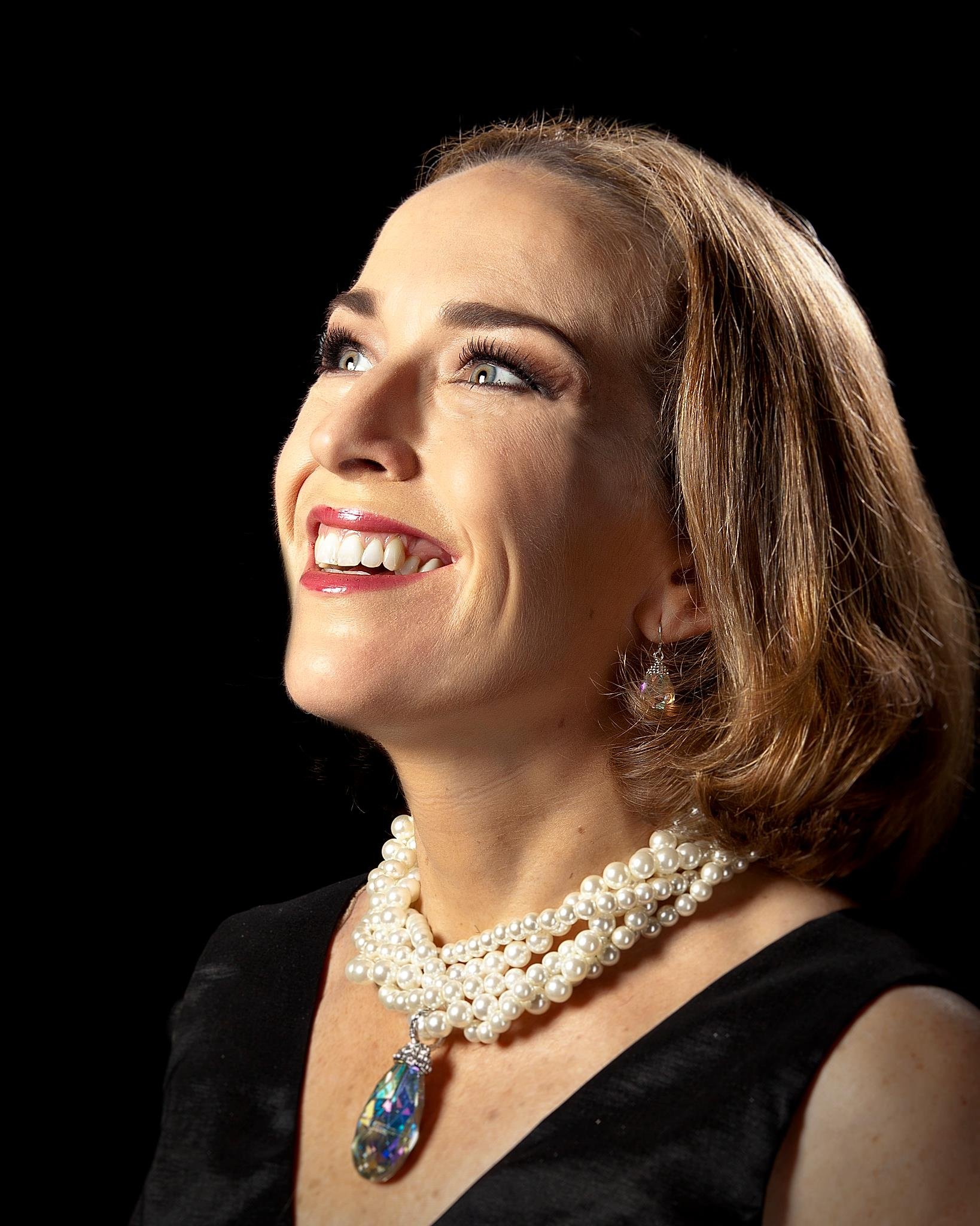Close-up of a smiling woman with light skin tone, wearing multiple pearl necklaces, a black top, and a colorful, teardrop-shaped pendant necklace, against a black background.