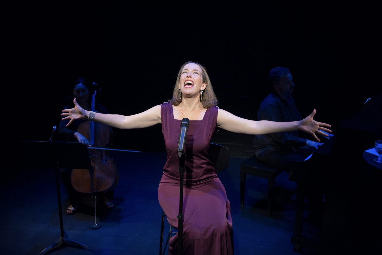 A woman in a purple dress singing passionately with her arms outstretched on stage, accompanied by a cellist and a pianist, both dressed in black, against a dark background.