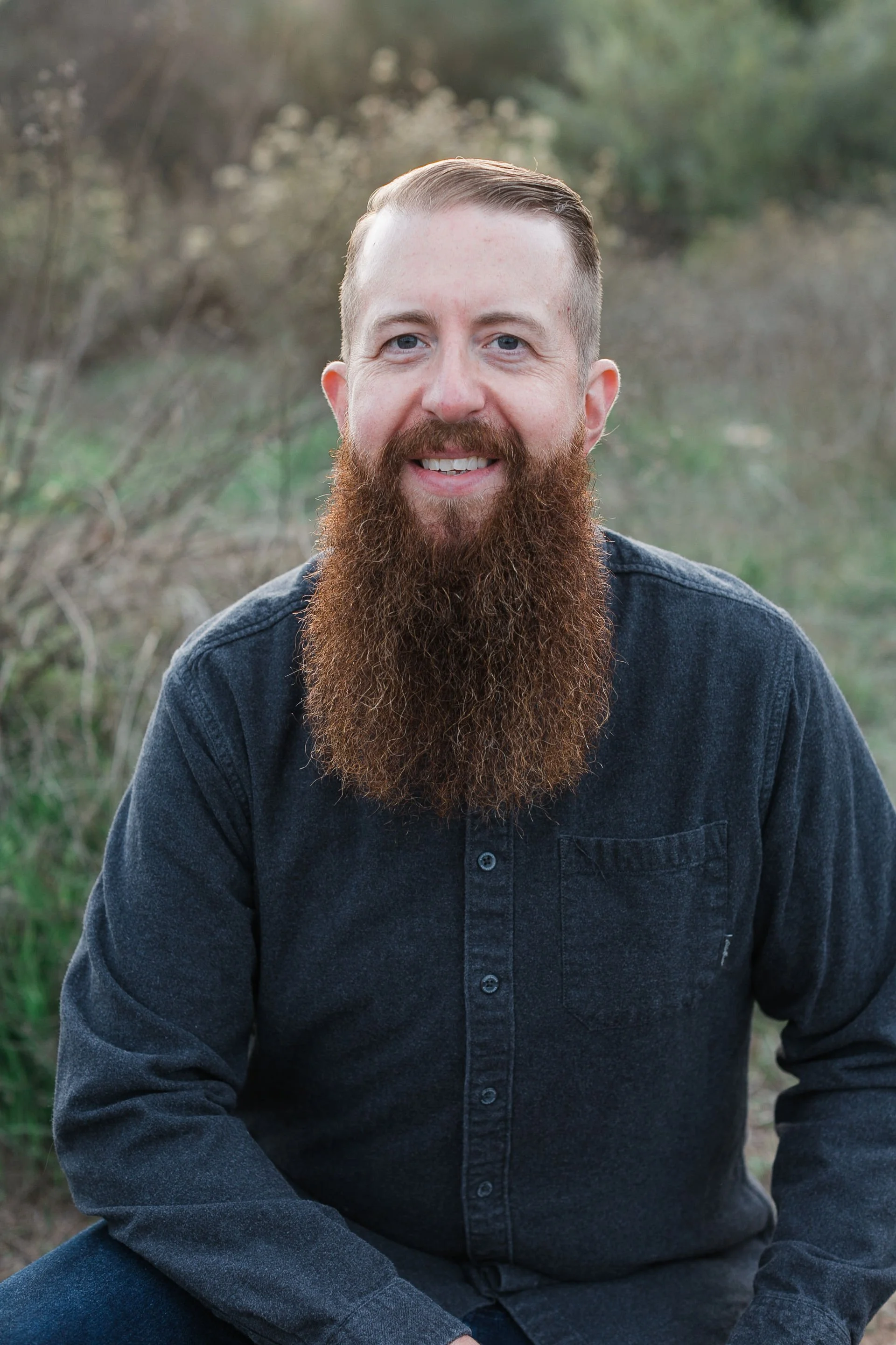 A man with blue eyes and a thick, reddish-brown beard smiling outdoors. He is wearing a dark grey shirt and sitting with his hands resting on his lap. The background is blurred with natural greenery.