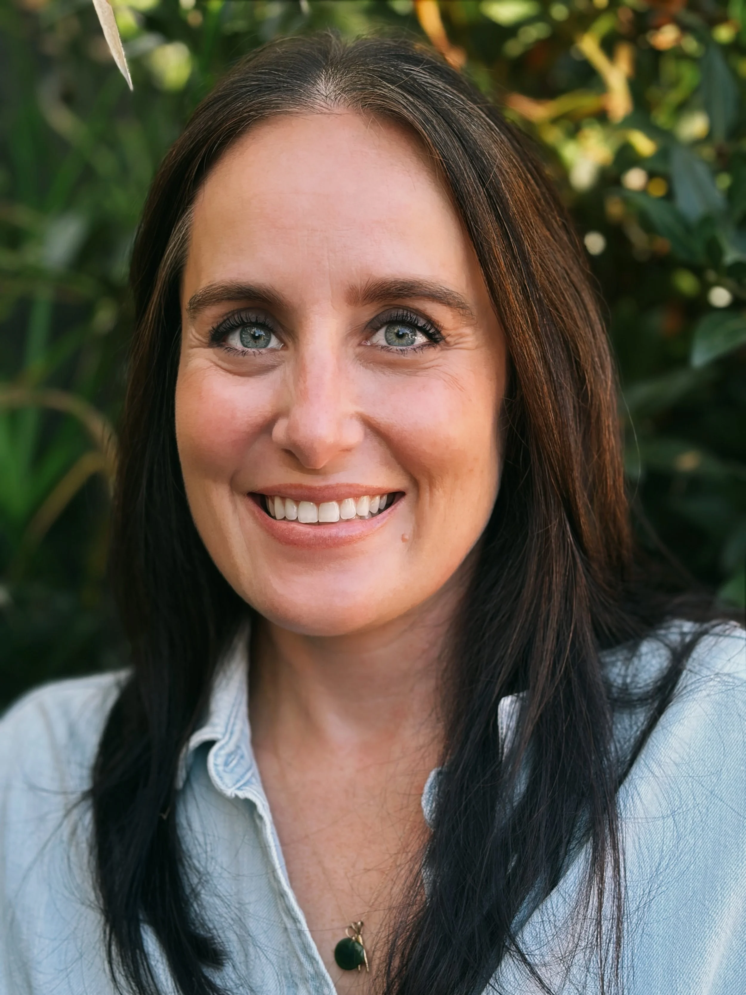 Close-up of a smiling woman with blue eyes and dark brown hair, wearing a light denim shirt and a necklace, outdoors in front of green foliage.