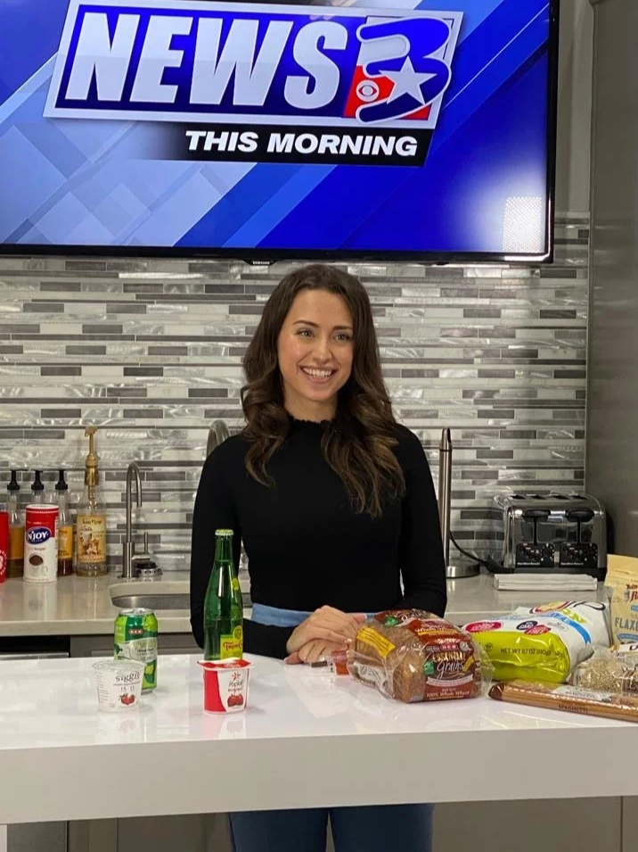 A woman standing behind a white table with various food and drink items, in a kitchen studio with a News channel logo on a screen above.