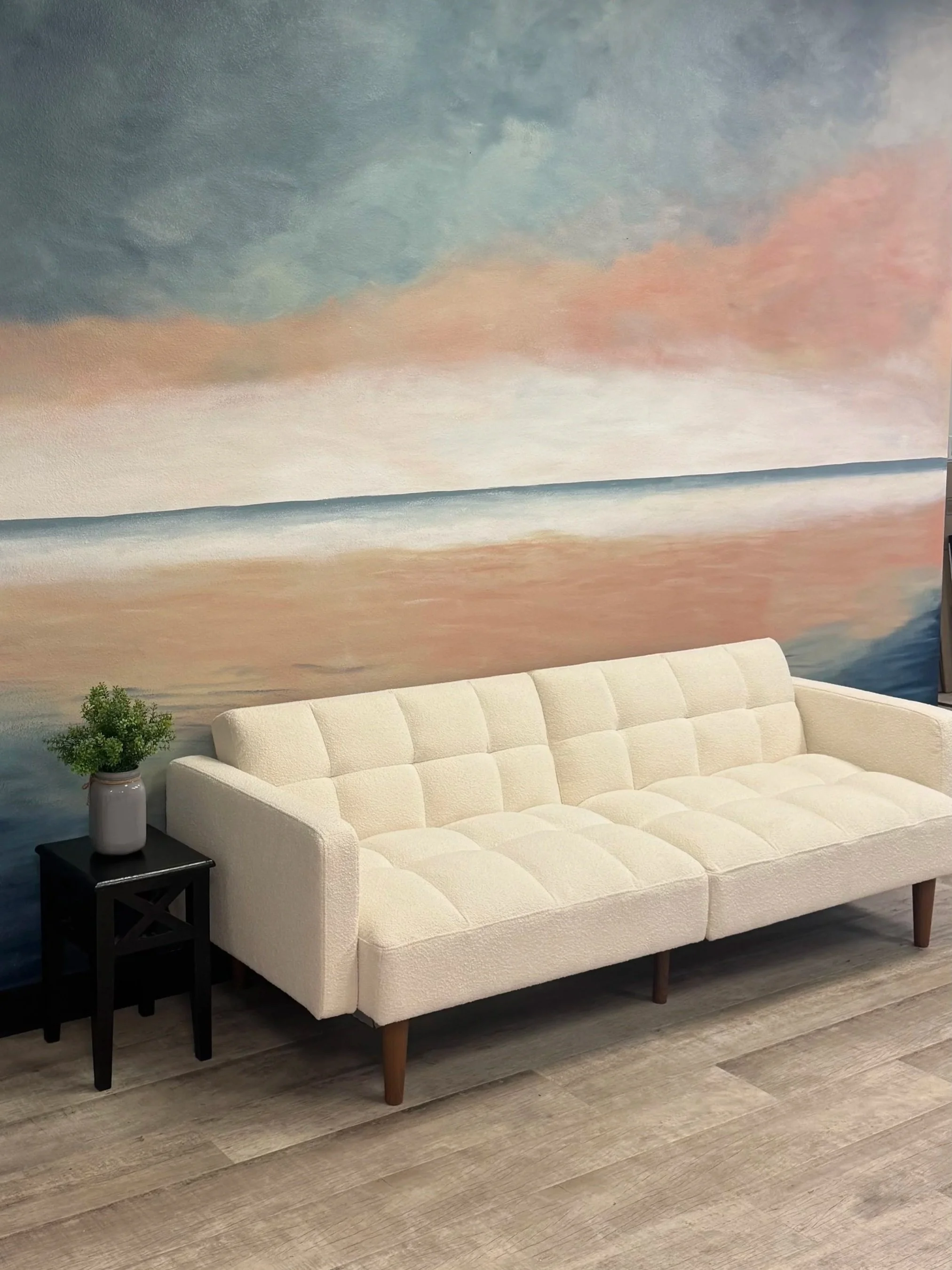 A white, mid-century modern sofa with tufted cushions and wooden legs in a living room, with a black side table and a potted plant on top, in front of a large abstract coastal-themed wall mural.