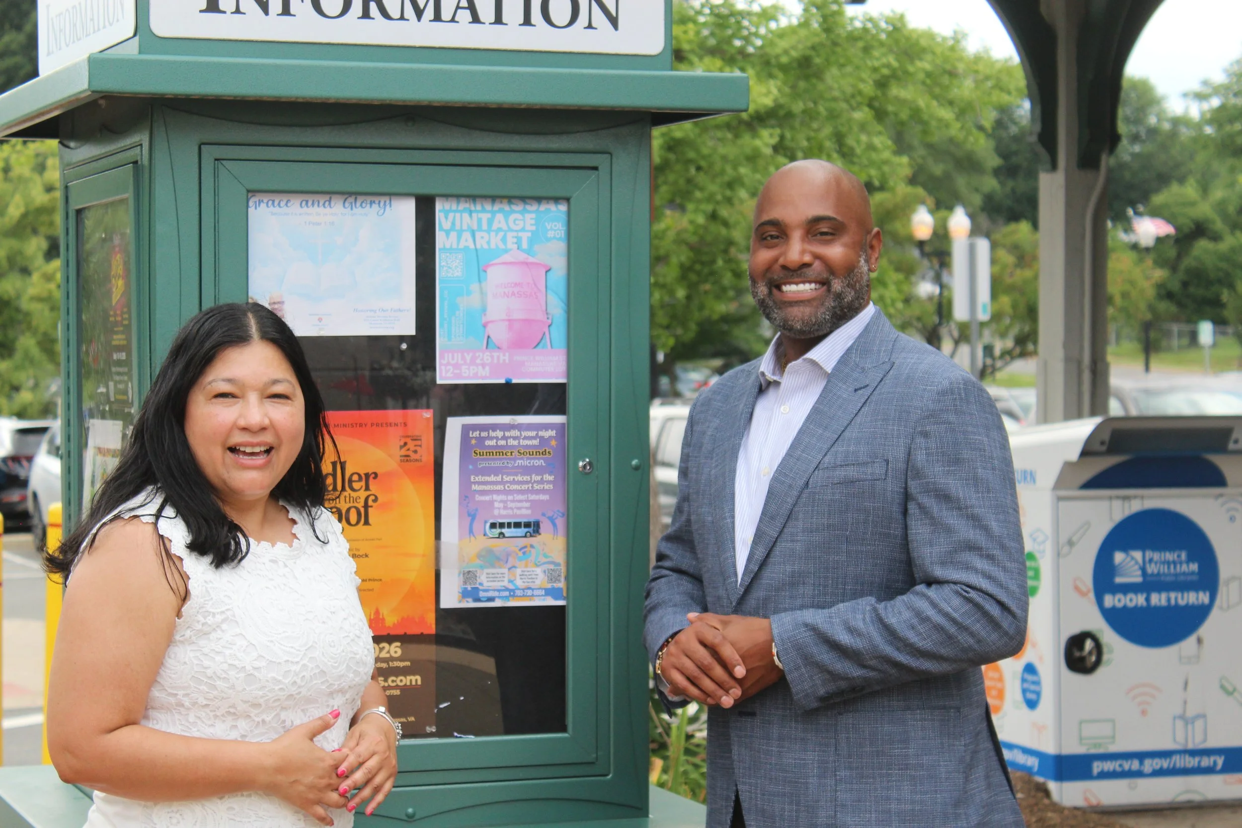 A woman and a man standing outdoors next to an information kiosk with various posters, with trees and street lights in the background.