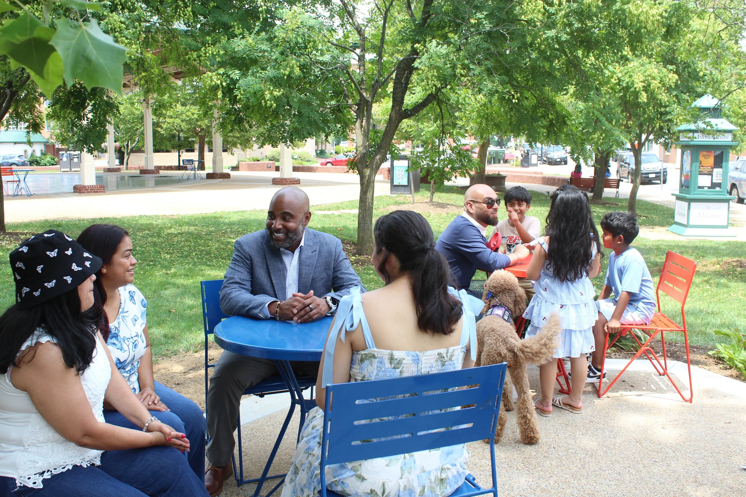 A diverse group of people, including adults and children, are sitting and standing in a park under green trees, engaging in conversation and enjoying a sunny day.